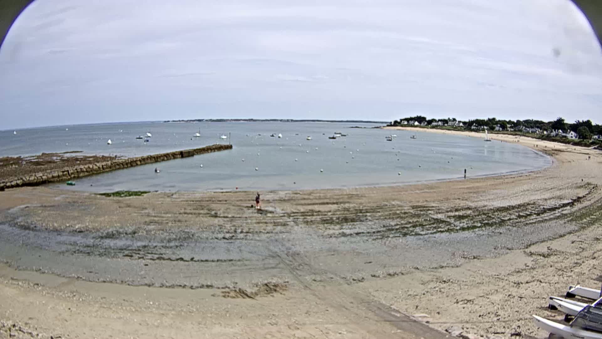 A sandy beach curves around a calm bay dotted with sailboats under a mostly cloudy sky.