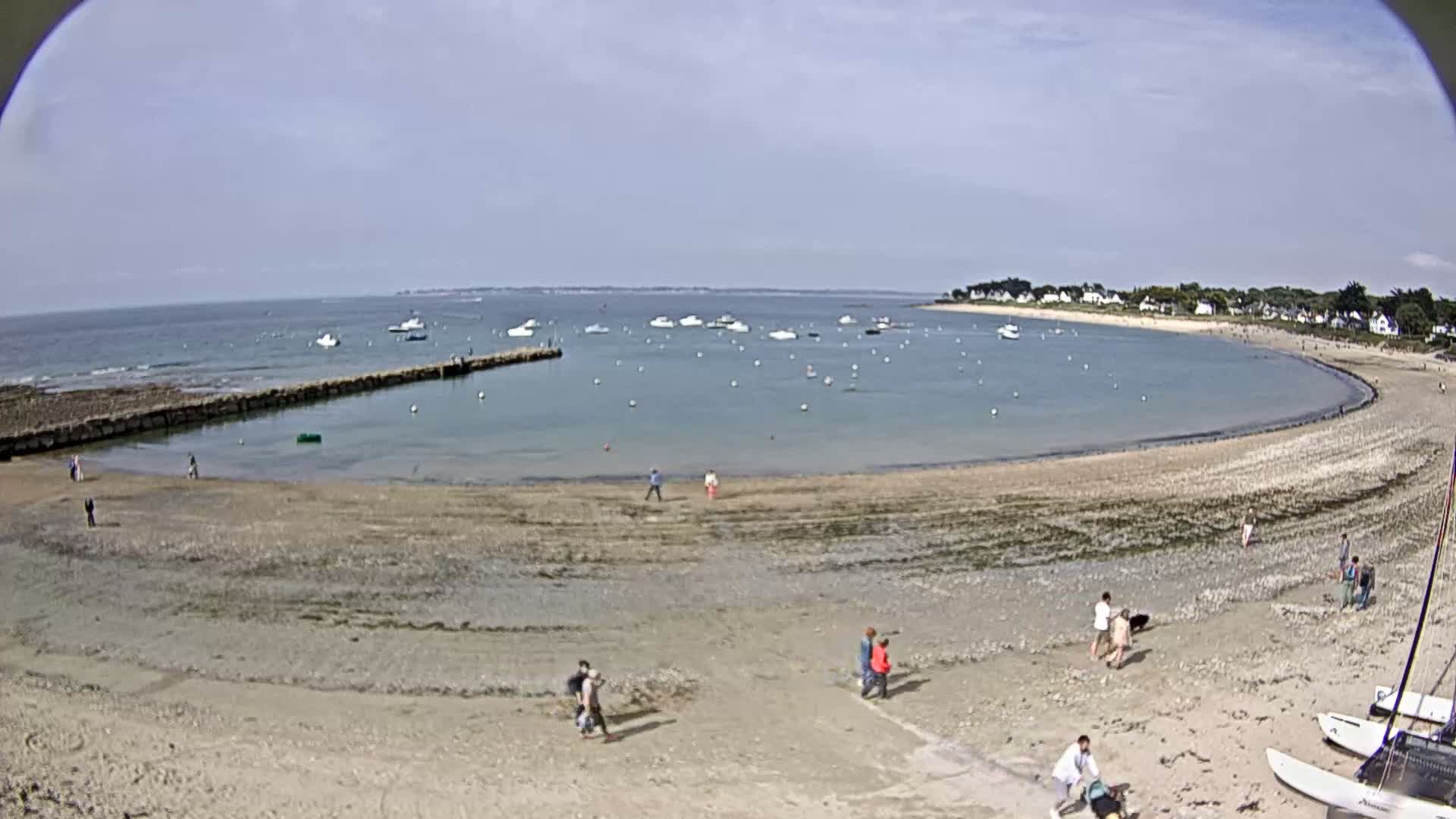 A sandy beach curves around a calm bay dotted with small boats under a mostly sunny sky.