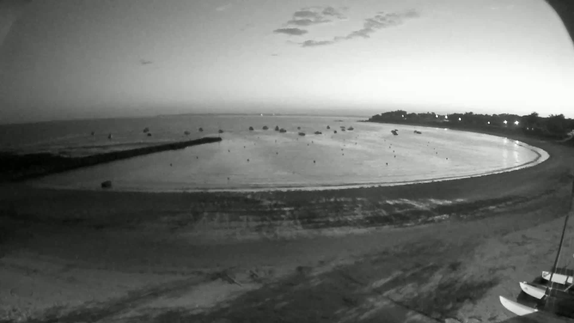 A calm, twilight bay with several boats at anchor is visible from a high vantage point, showing a sandy beach and a breakwater.