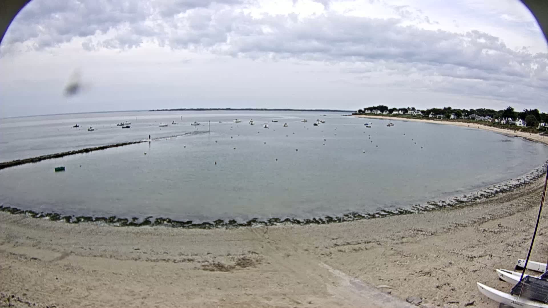 A calm bay with several boats is partially enclosed by a sandy beach under a cloudy sky.