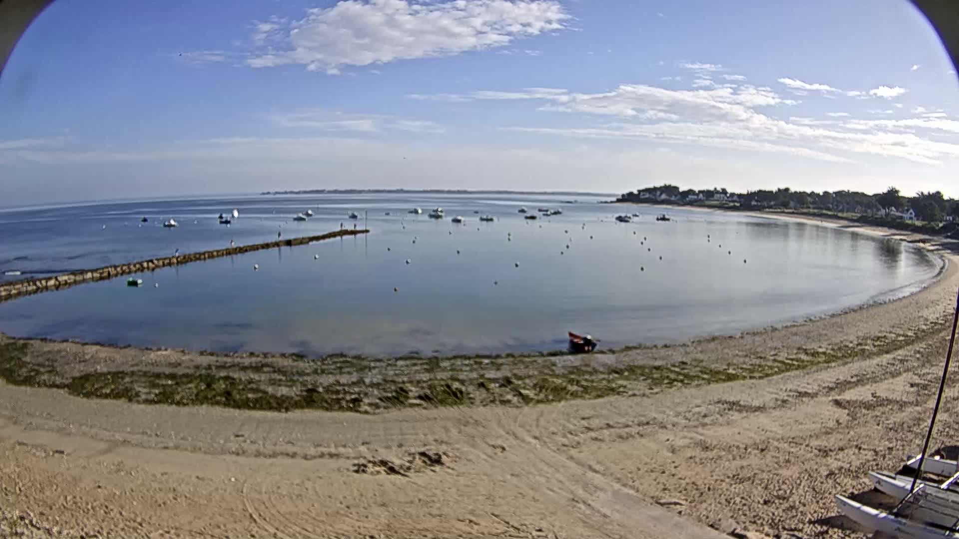 A sandy beach curves around a calm bay dotted with small boats under a mostly sunny sky.