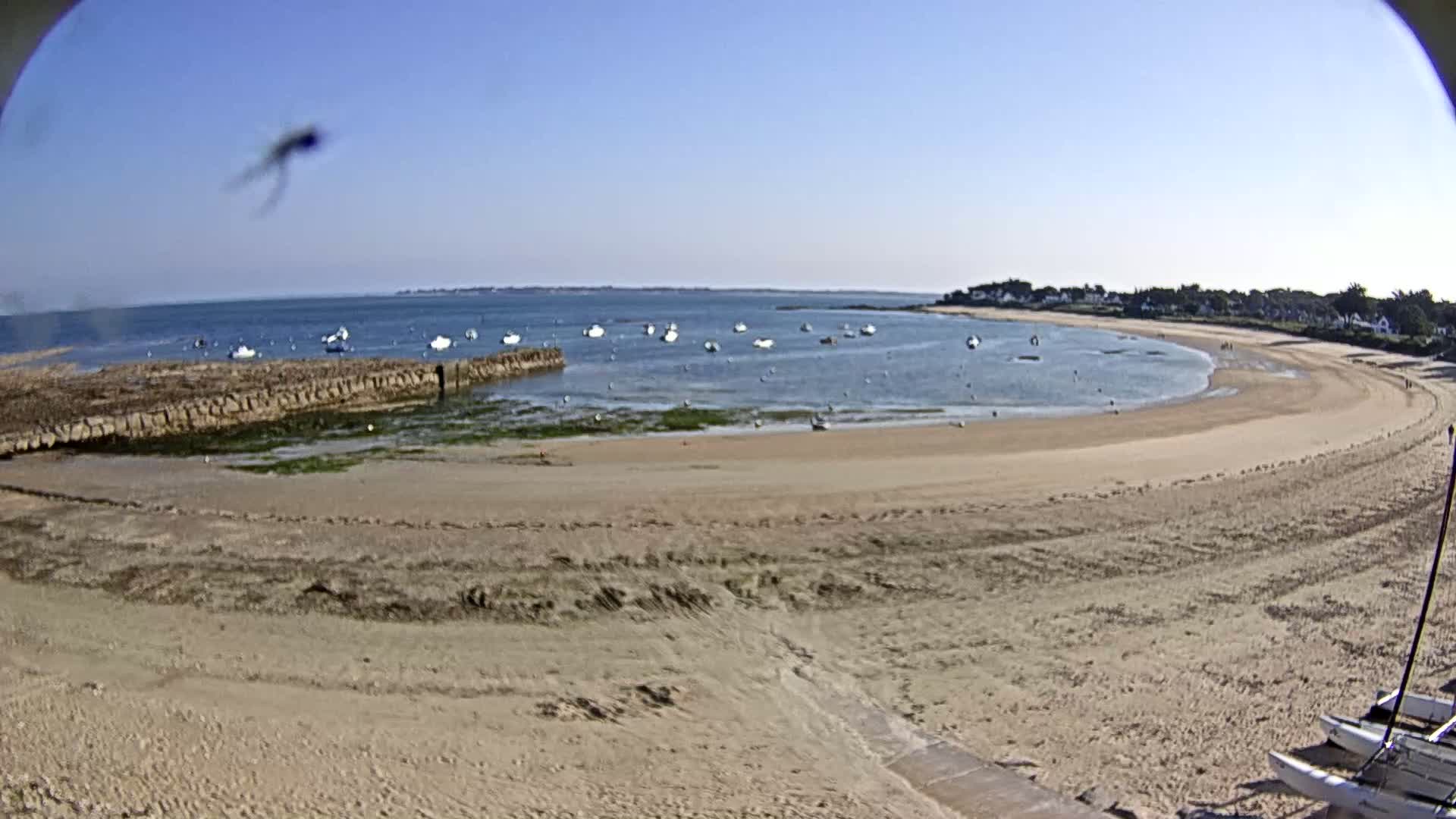 A sandy beach curves around a calm bay with several small boats anchored in the water under a clear blue sky.