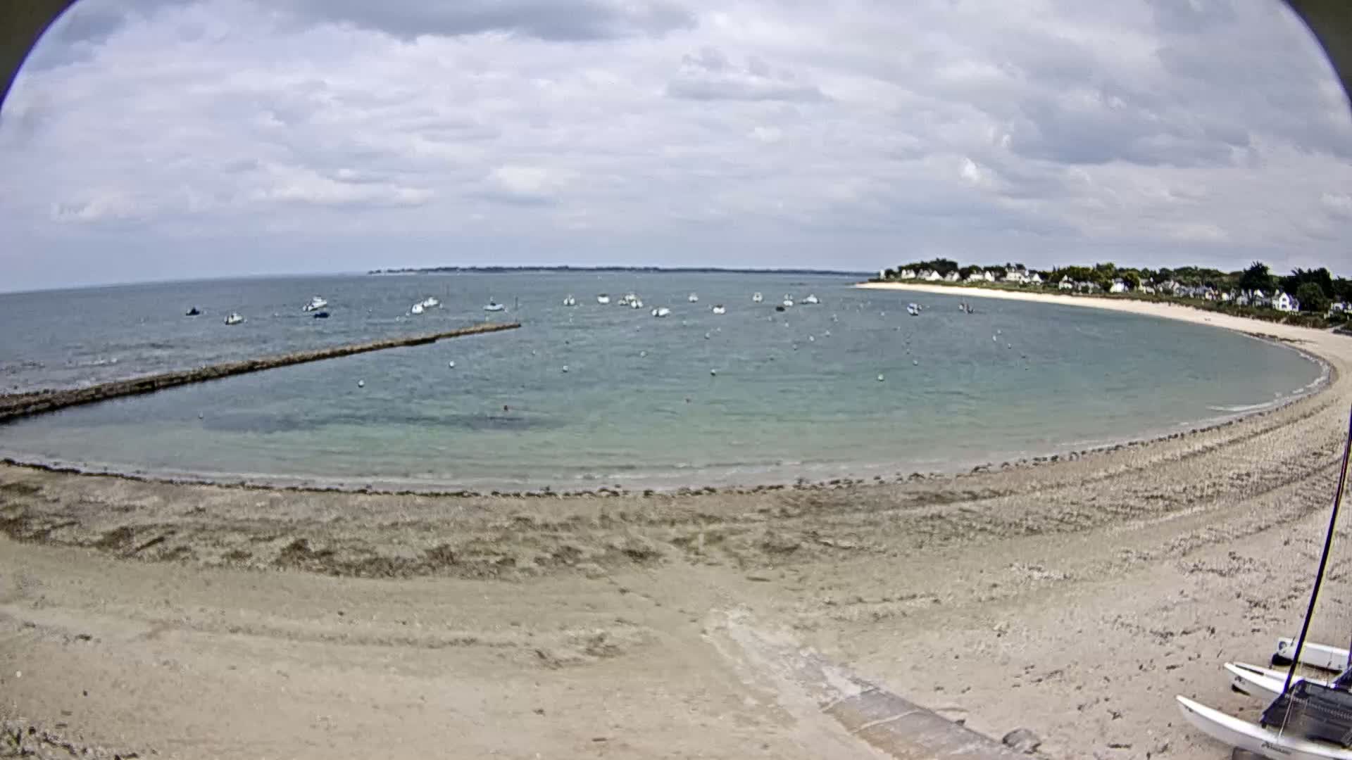 A sandy beach curves around a calm, light-green bay dotted with small boats under a cloudy sky.