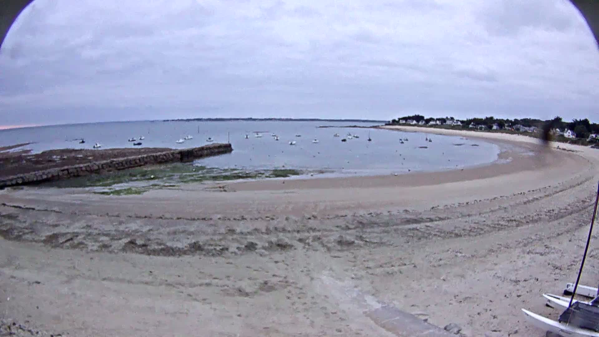 A sandy beach curves around a calm bay with several small boats anchored, under an overcast sky.