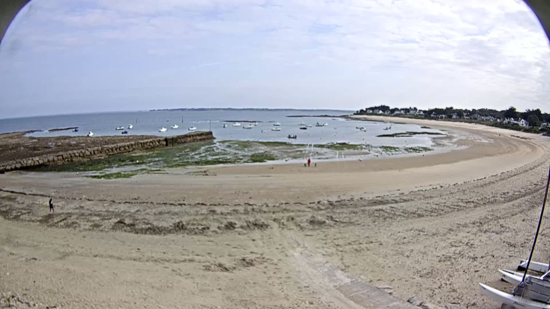 A sandy beach curves around a calm bay with several small boats, under a partly cloudy sky.