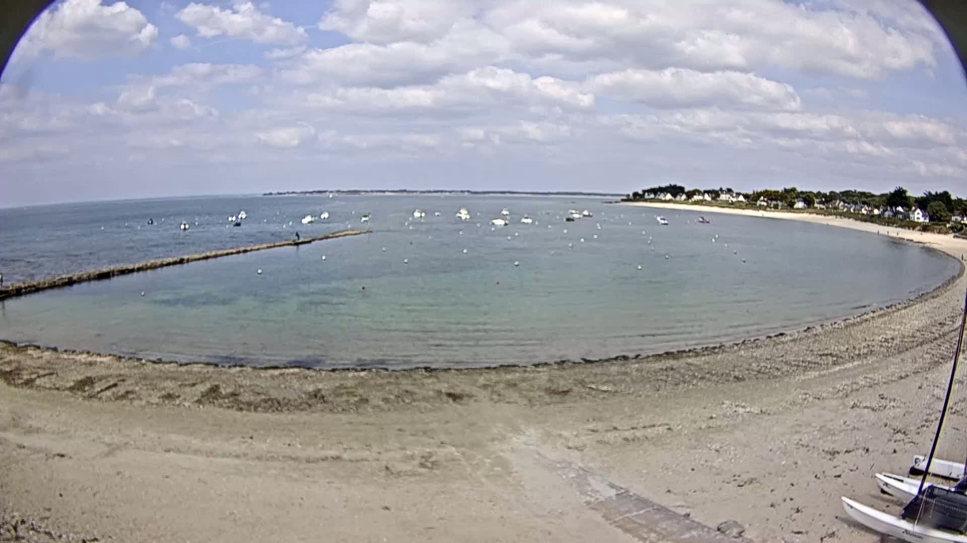 A sandy beach curves around a calm, shallow bay dotted with small boats under a partly cloudy sky.