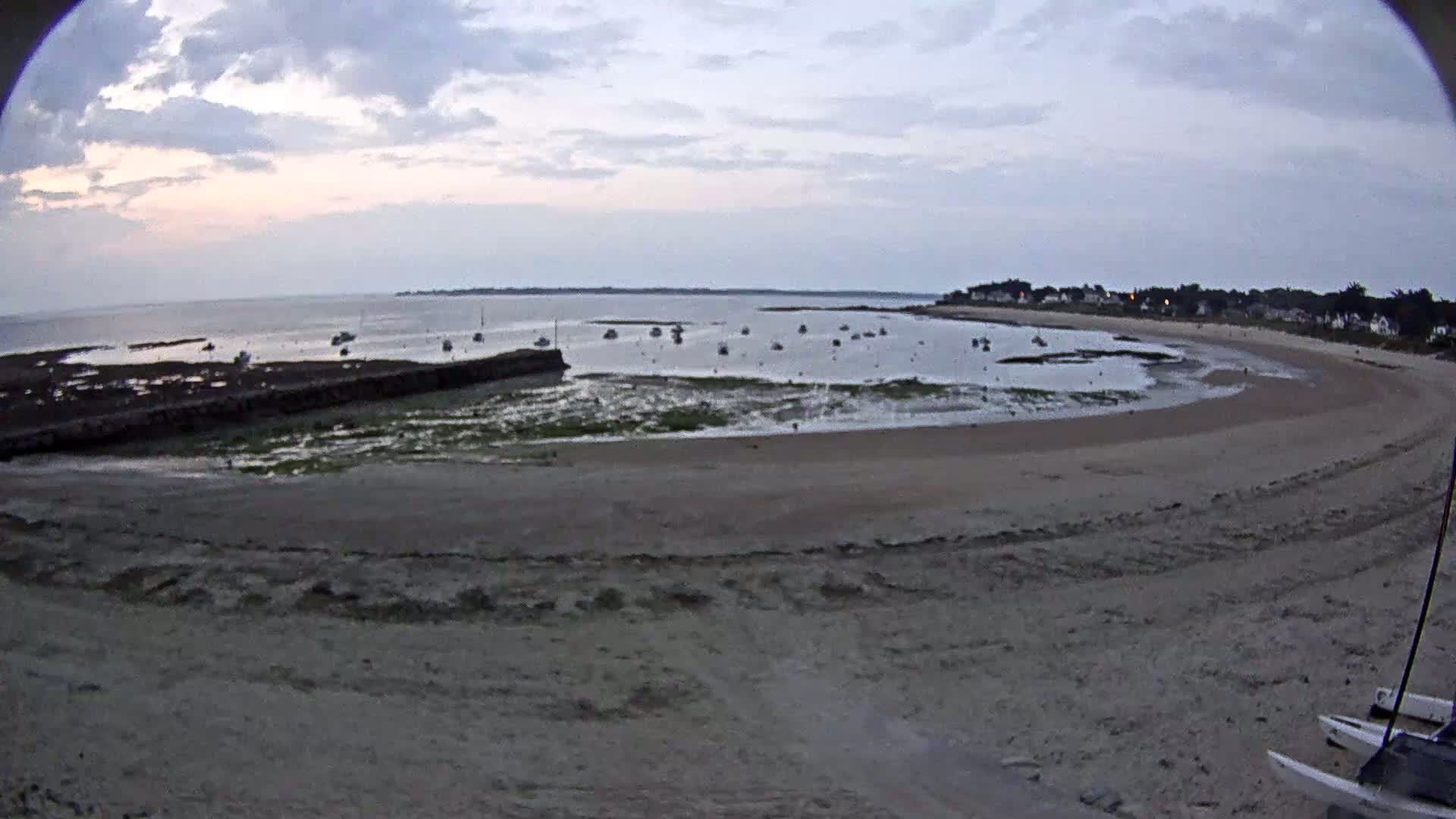 A sandy beach curves around a calm bay with numerous small boats at anchor under a cloudy, dusky sky.