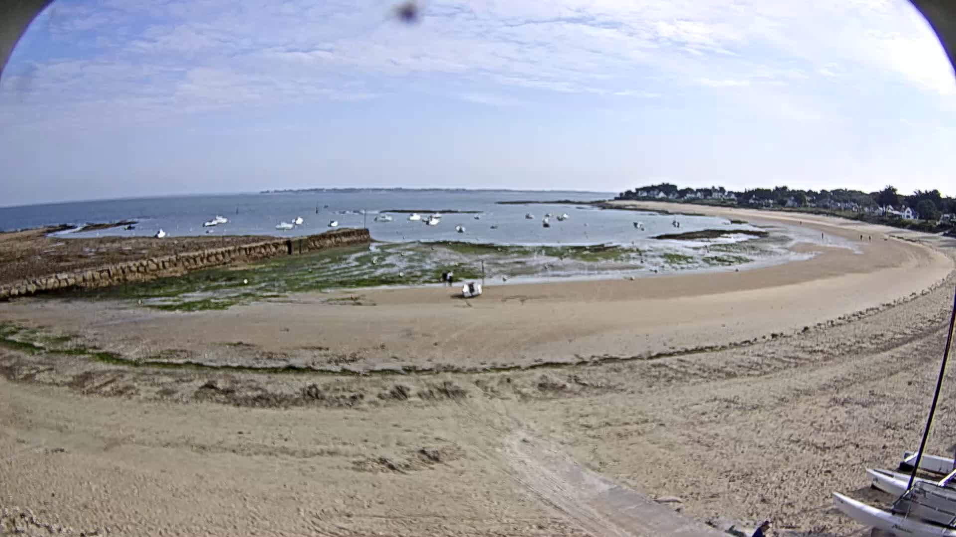 A sandy beach curves around a calm bay with several small boats anchored in shallow water under a partly cloudy sky.