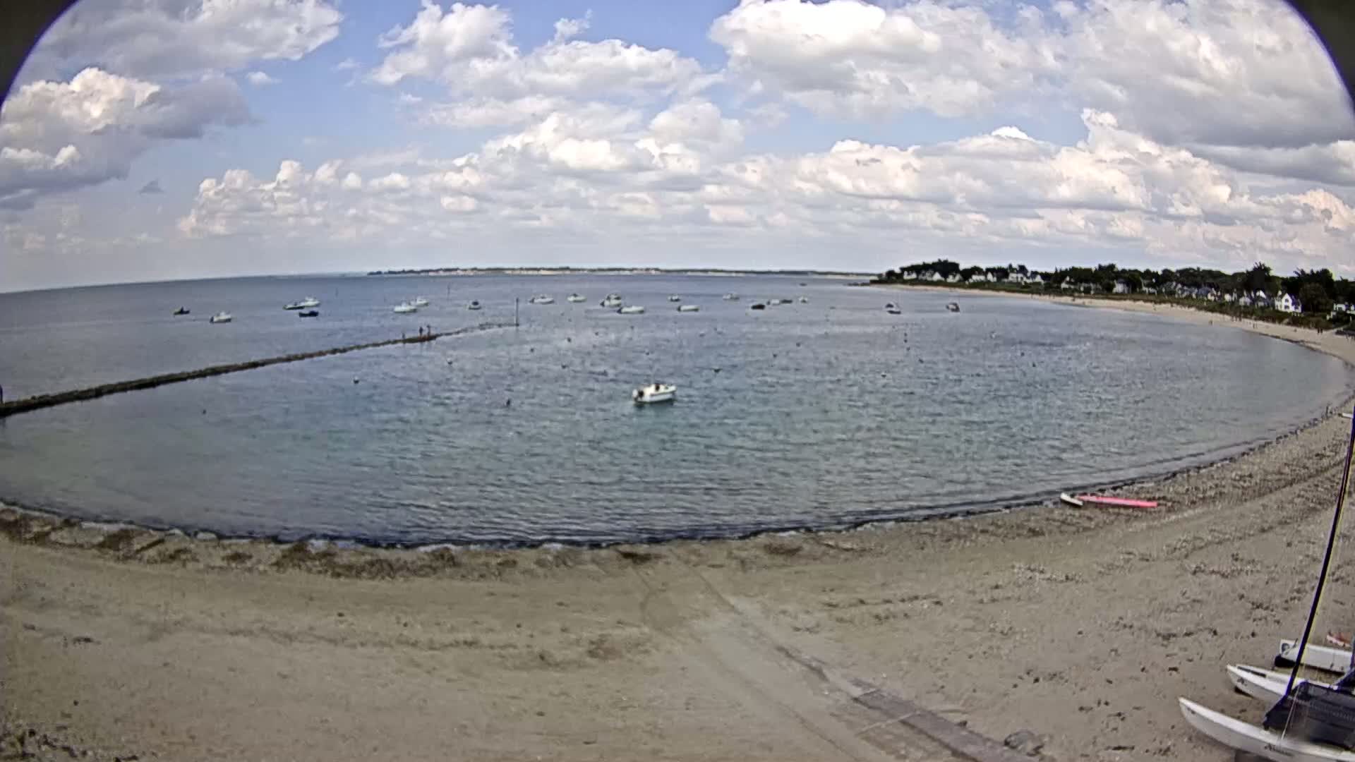 A sandy beach curves around a calm bay with numerous small boats under a partly cloudy sky.