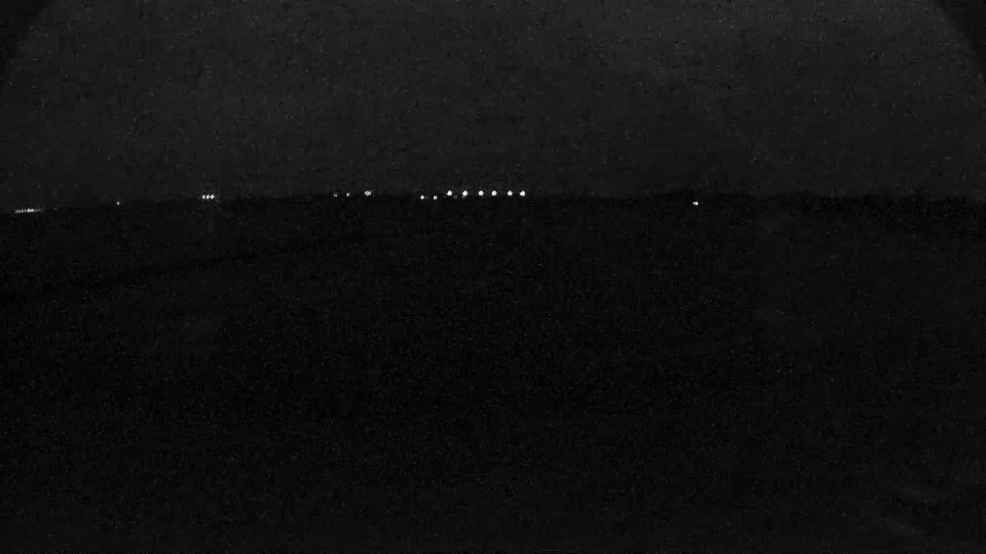 A dark, nighttime scene shows a line of distant lights on a flat horizon.