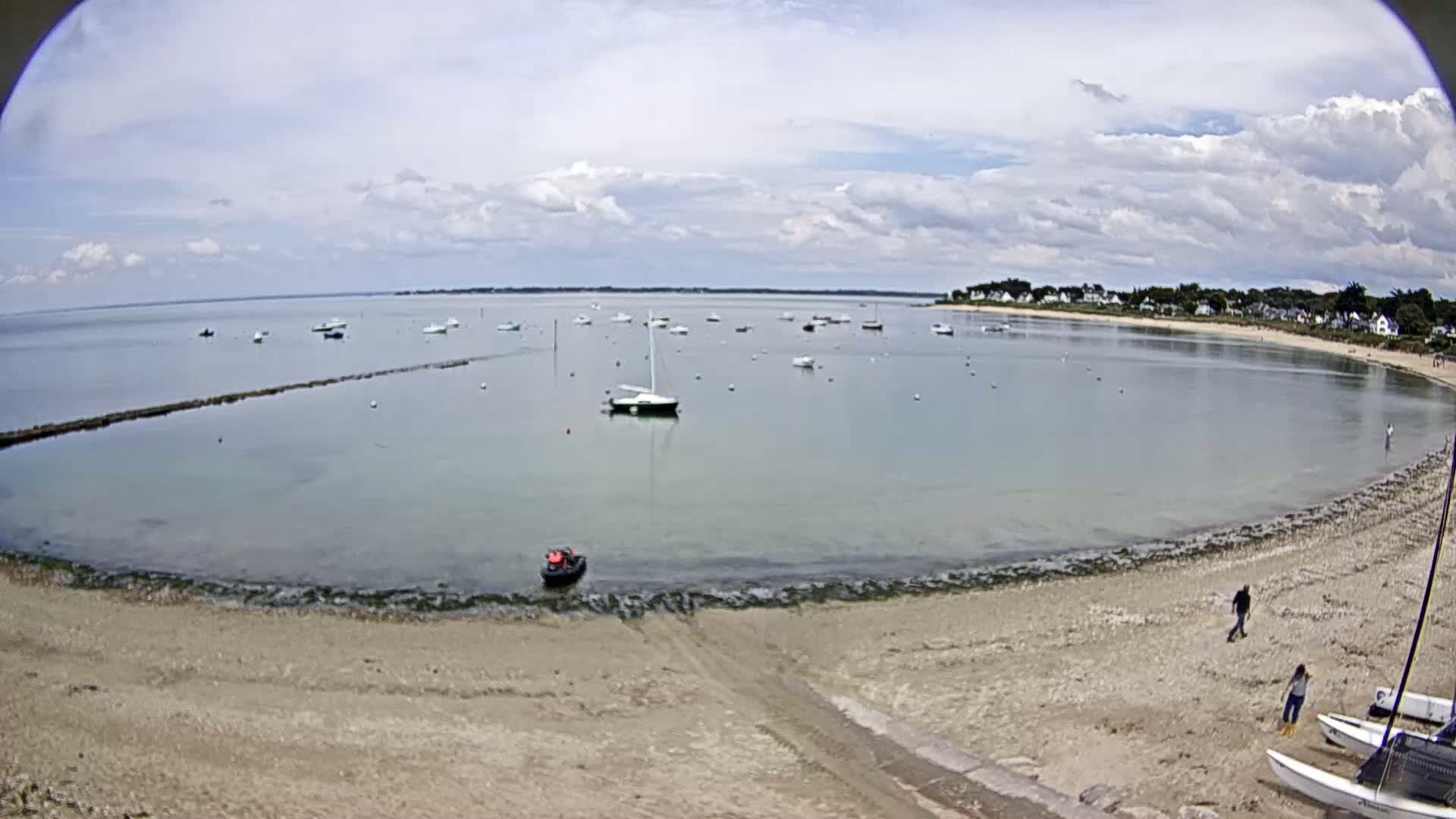 A sandy beach curves around a calm bay with numerous boats at anchor under a partly cloudy sky.