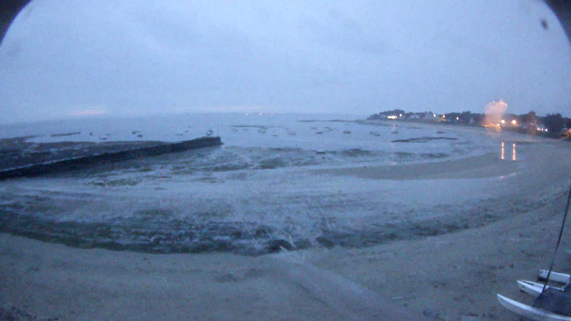 Small waves lap a sandy beach at dusk, with several boats visible in the calm water beyond a low seawall.