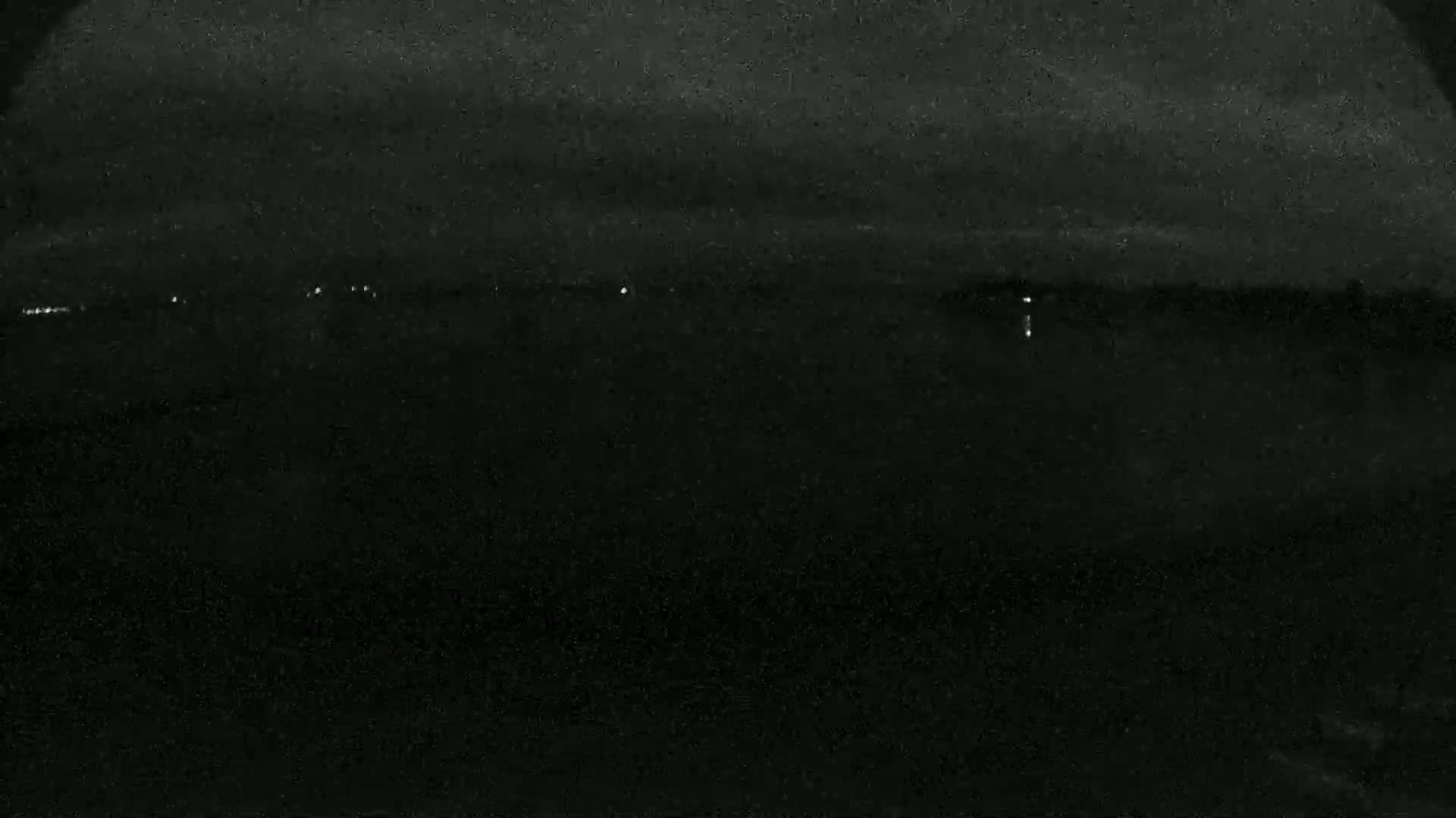 A dark, nighttime view of a flat, dark landscape with a few faint lights visible in the distance under a cloudy sky.
