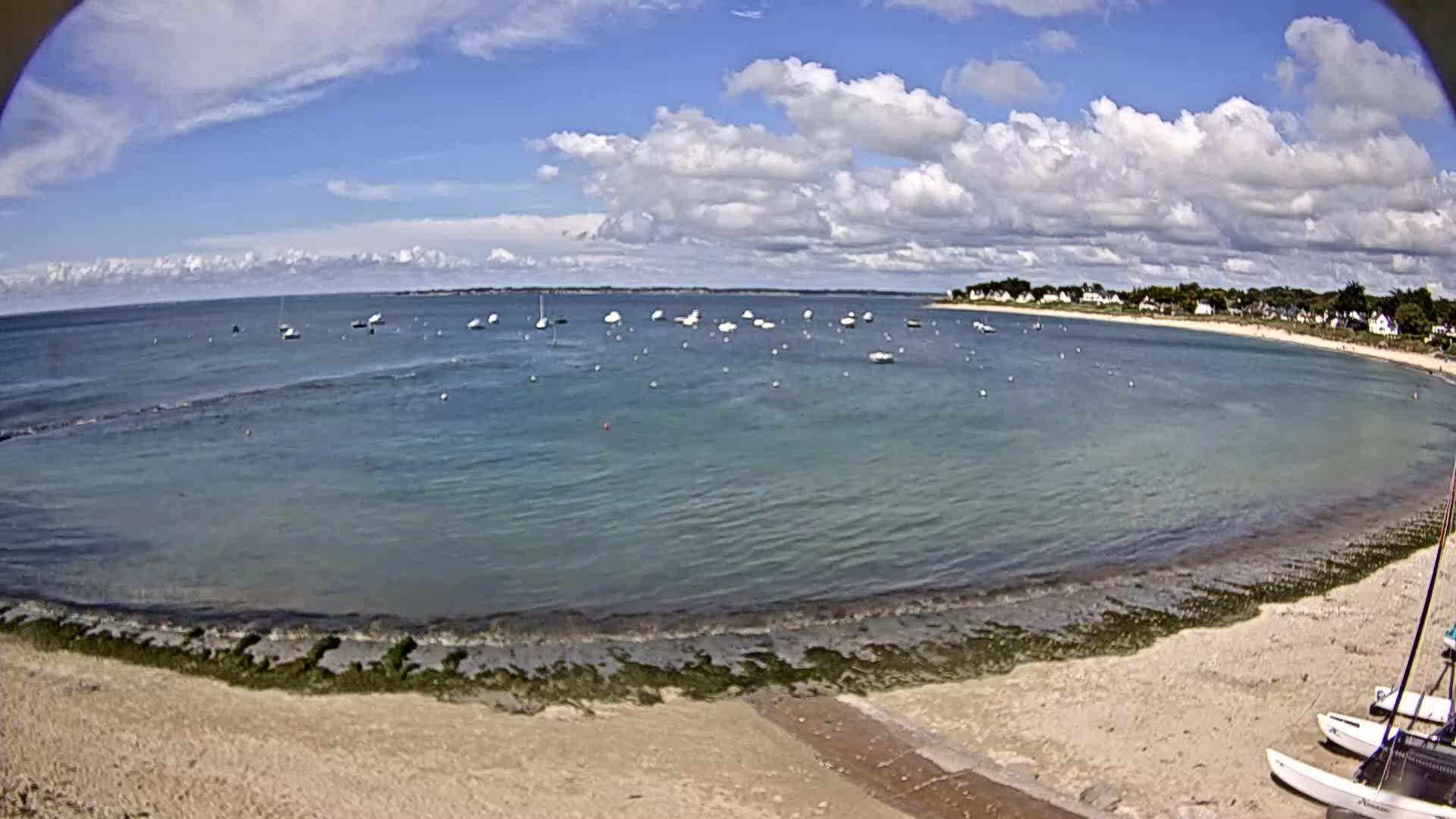 A partly cloudy day shows a calm bay with numerous small sailboats anchored, a sandy beach with seaweed at the water's edge, and a small village in the distance.