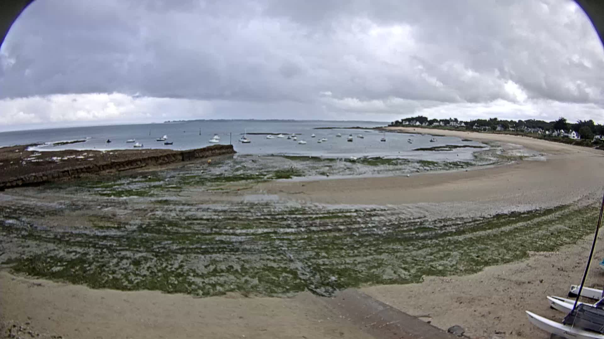 A wide, sandy beach curves around a calm bay under a cloudy sky, with several boats anchored in the water and seaweed visible along the shoreline.