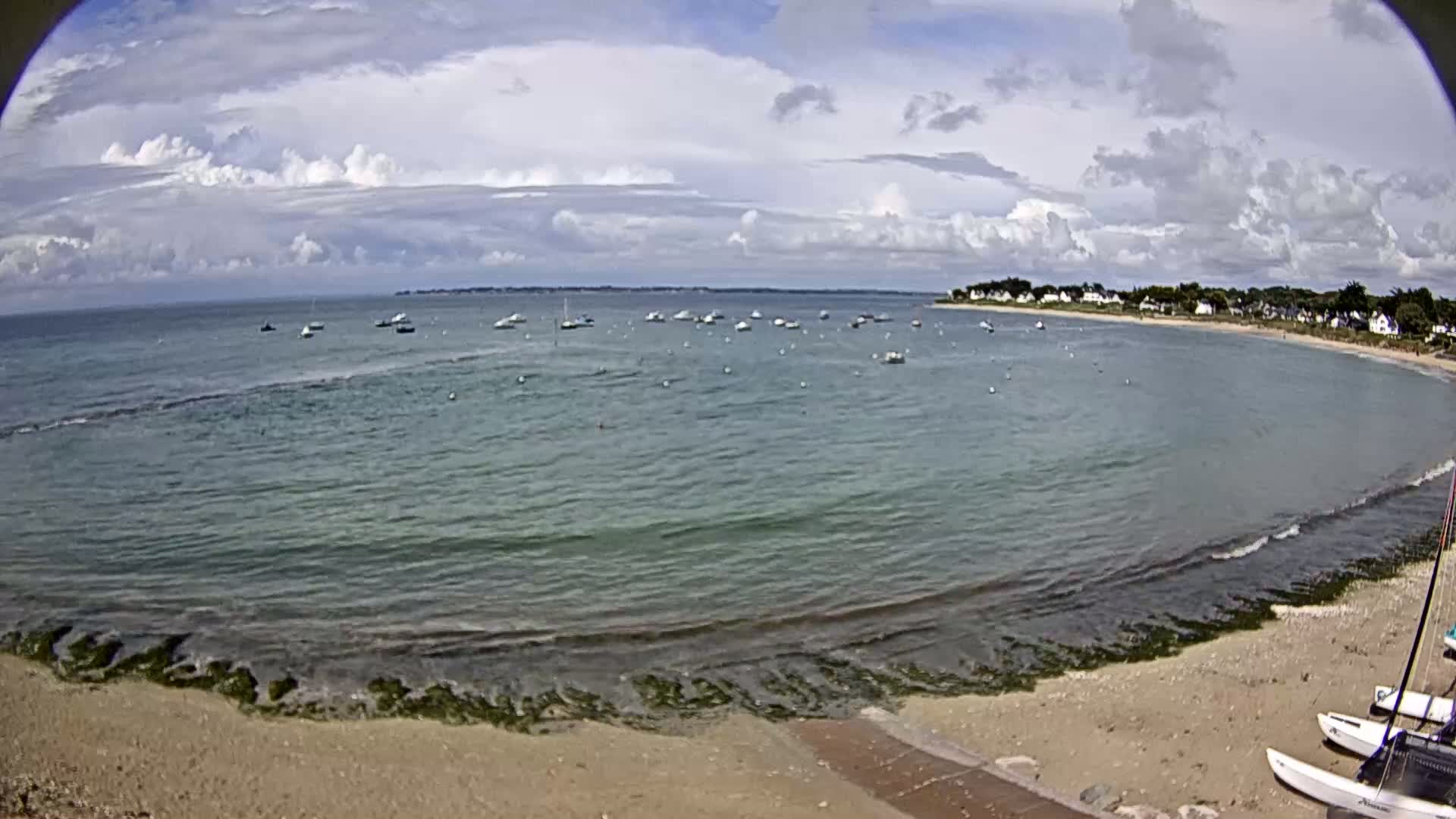 A partly cloudy day shows a sandy beach with several sailboats anchored in calm, shallow water near a small village.