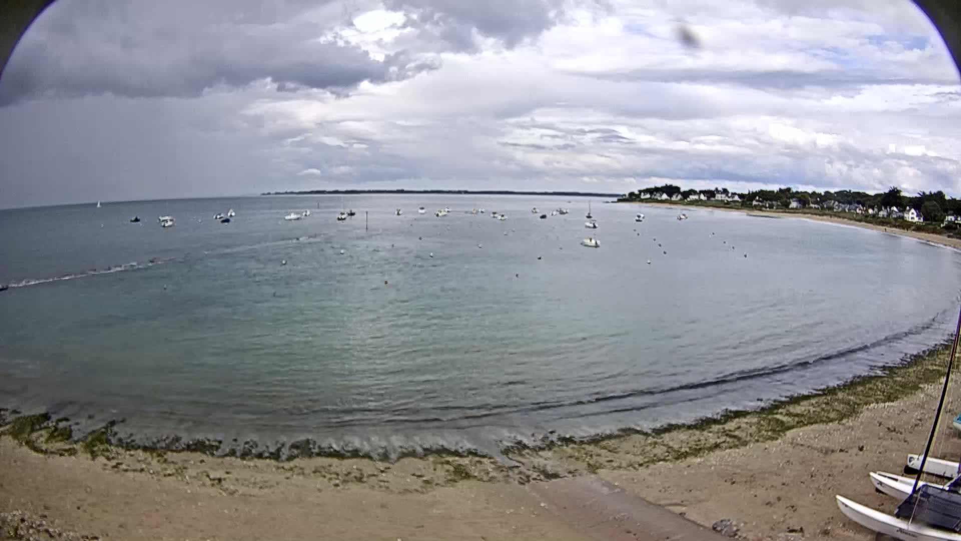 A calm bay with numerous small boats is visible from a sandy beach under a mostly cloudy sky.