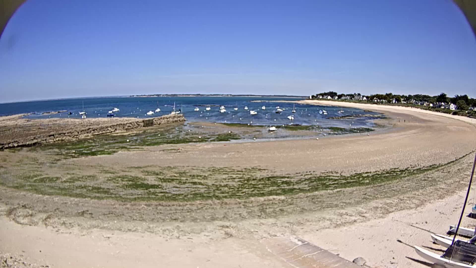 A sandy beach curves around a calm bay, where numerous small boats are anchored in shallow water under a clear blue sky.