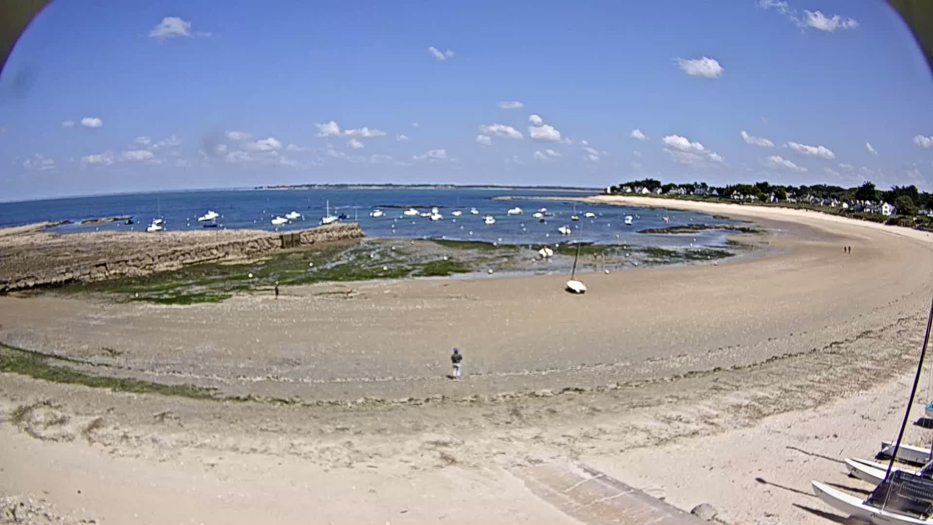 A sandy beach with several small boats in the water, under a mostly sunny sky, shows a rocky shoreline and a few people walking on the sand.
