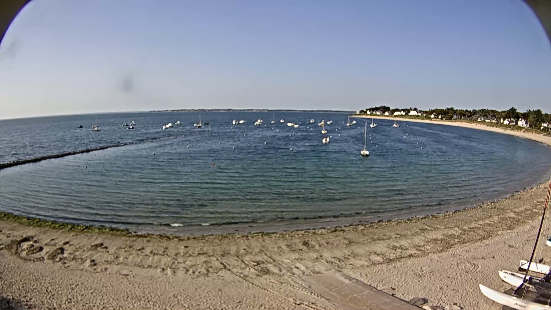 A sandy beach curves around a calm bay dotted with numerous small boats under a clear blue sky.