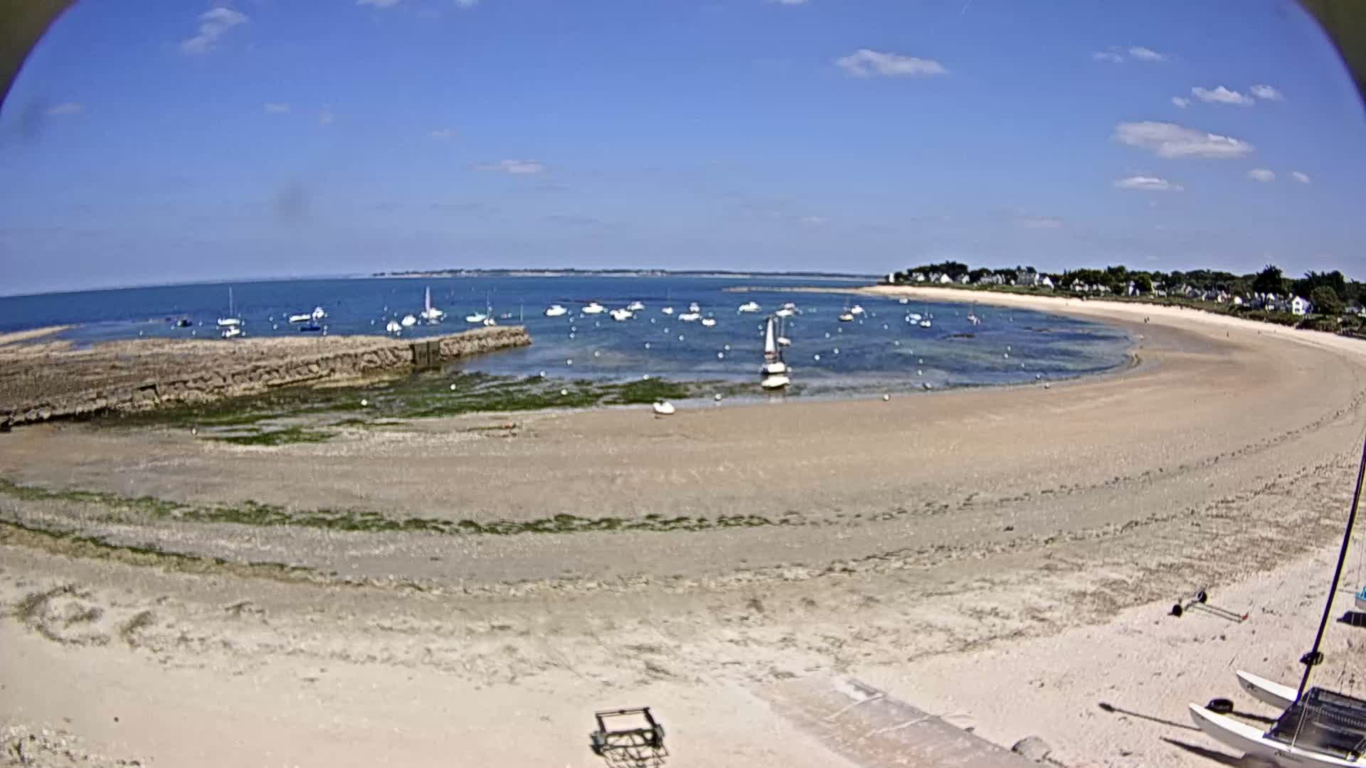 A sandy beach curves around a calm bay with numerous small sailboats anchored, under a clear blue sky.