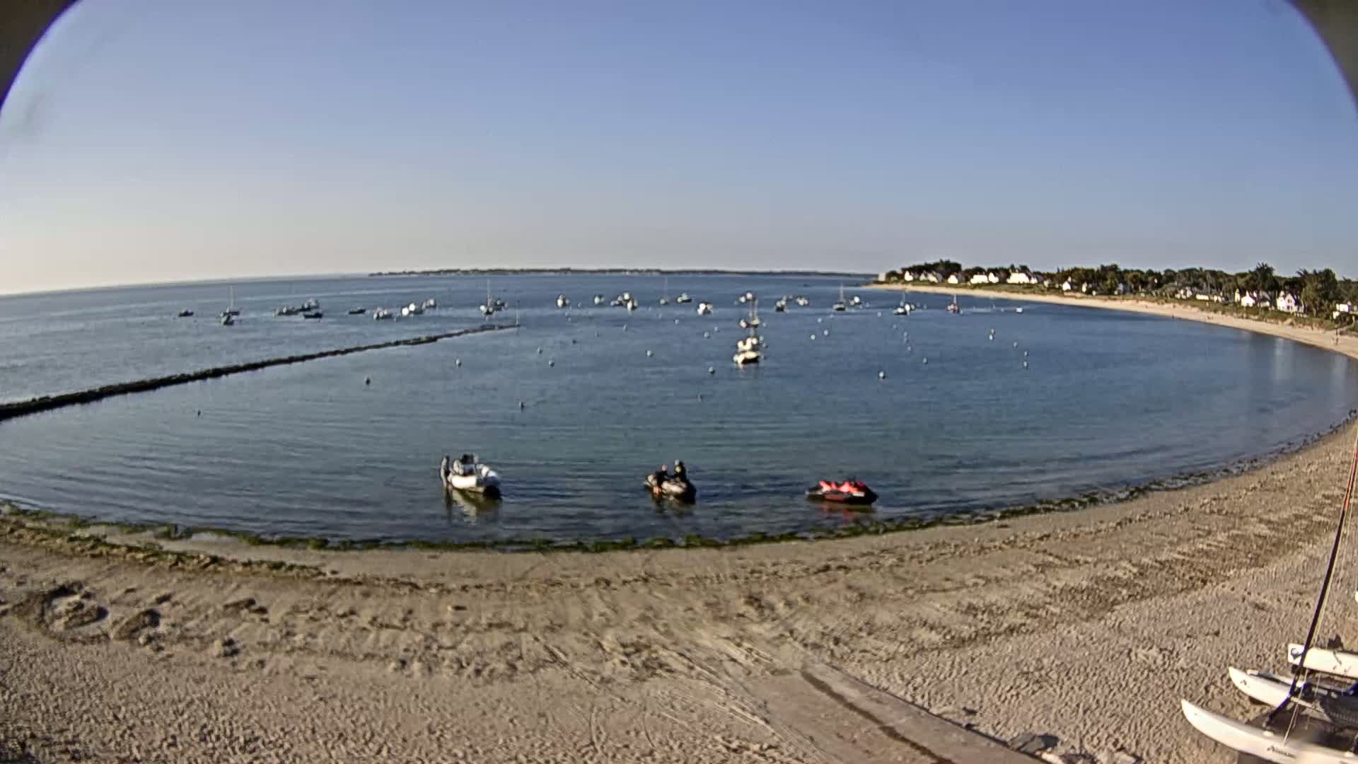 A sandy beach curves around a calm, blue bay dotted with small boats and jet skis under a clear, sunny sky.