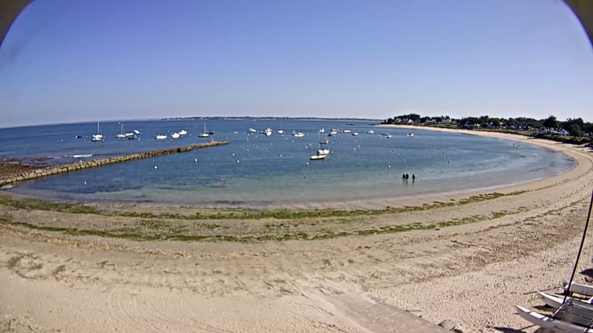 A sandy beach curves around a calm, blue bay dotted with sailboats under a clear blue sky.