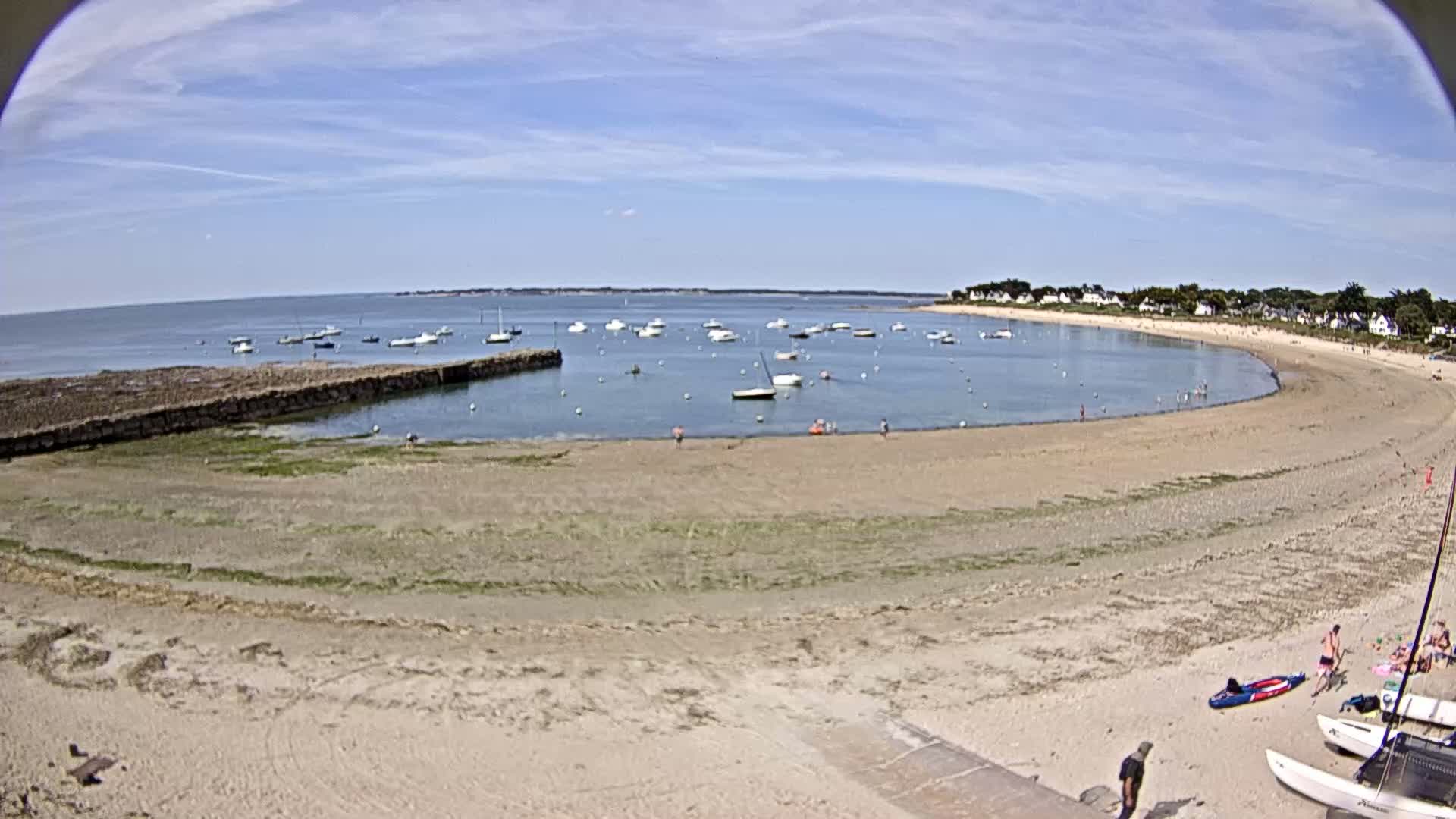 A sandy beach curves around a calm bay filled with small boats under a mostly sunny sky.
