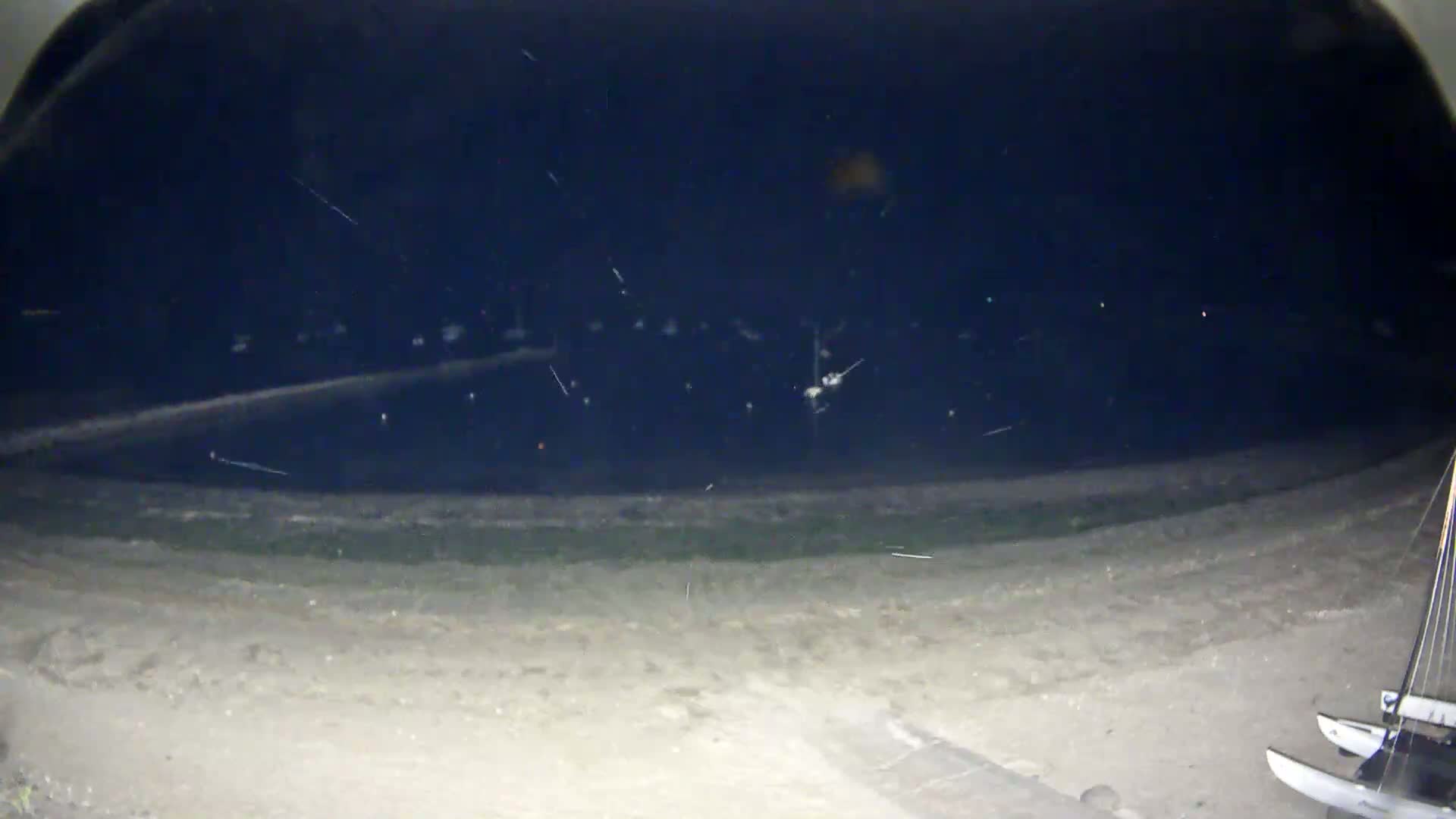 At night, a sandy shore leads to a dark body of water with numerous small lights and what appears to be rain or snow falling.