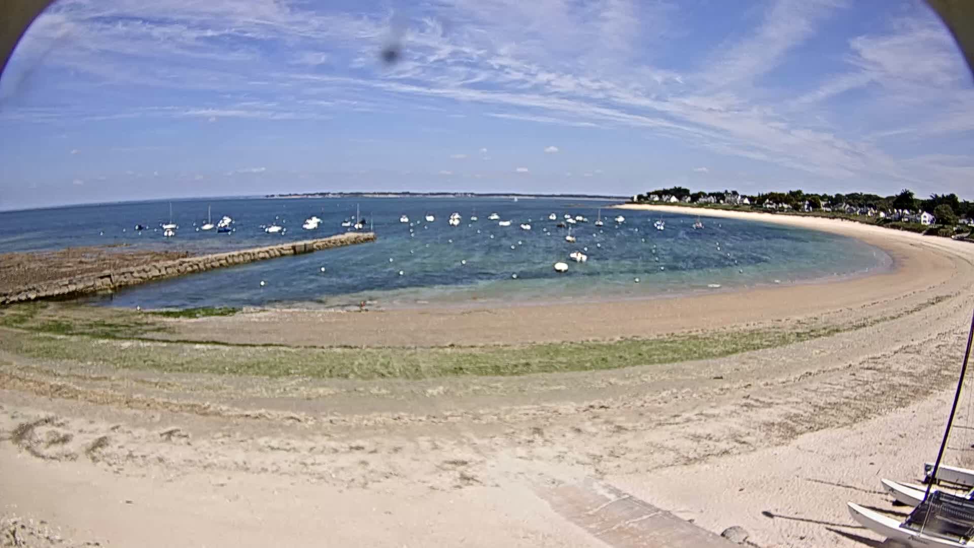 A sandy beach curves around a calm bay filled with numerous small boats under a mostly sunny sky.
