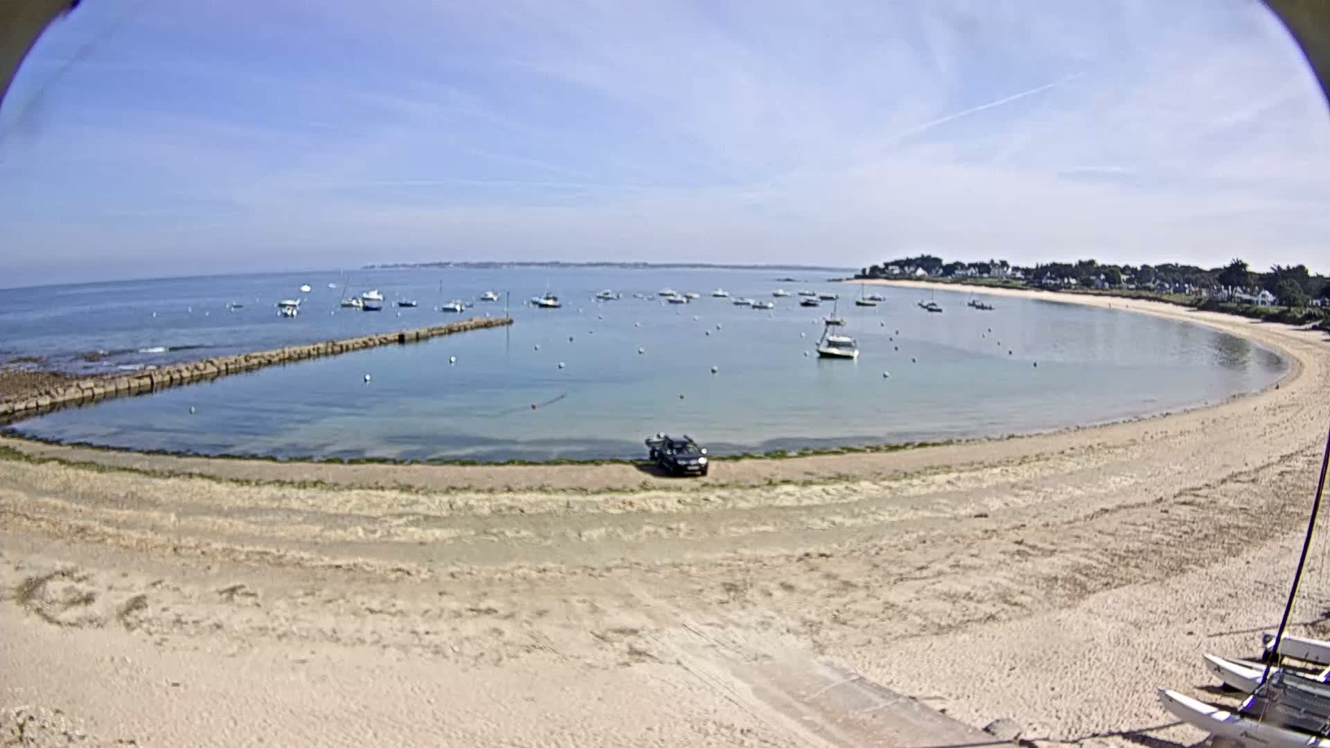A sandy cove with numerous boats in calm water under a clear blue sky is viewed from a slight elevation.