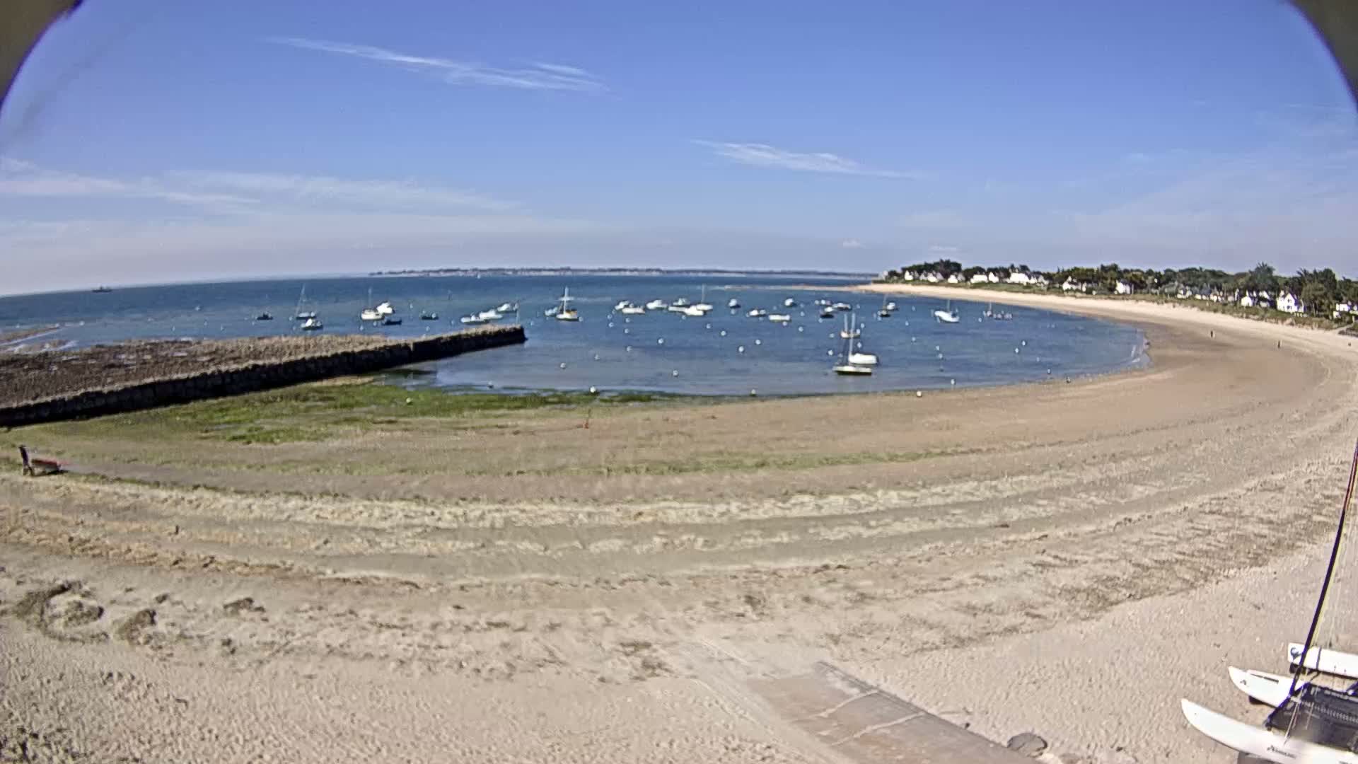 A sandy beach curves around a calm bay dotted with sailboats under a clear blue sky.