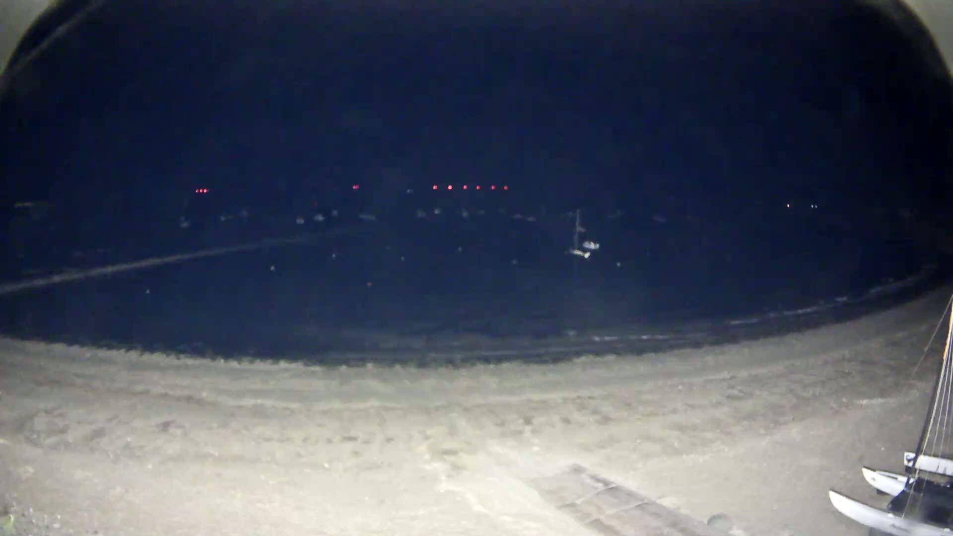A nighttime view from a beach shows a dark body of water with a few small lights in the distance, and two catamarans on the sand in the foreground.