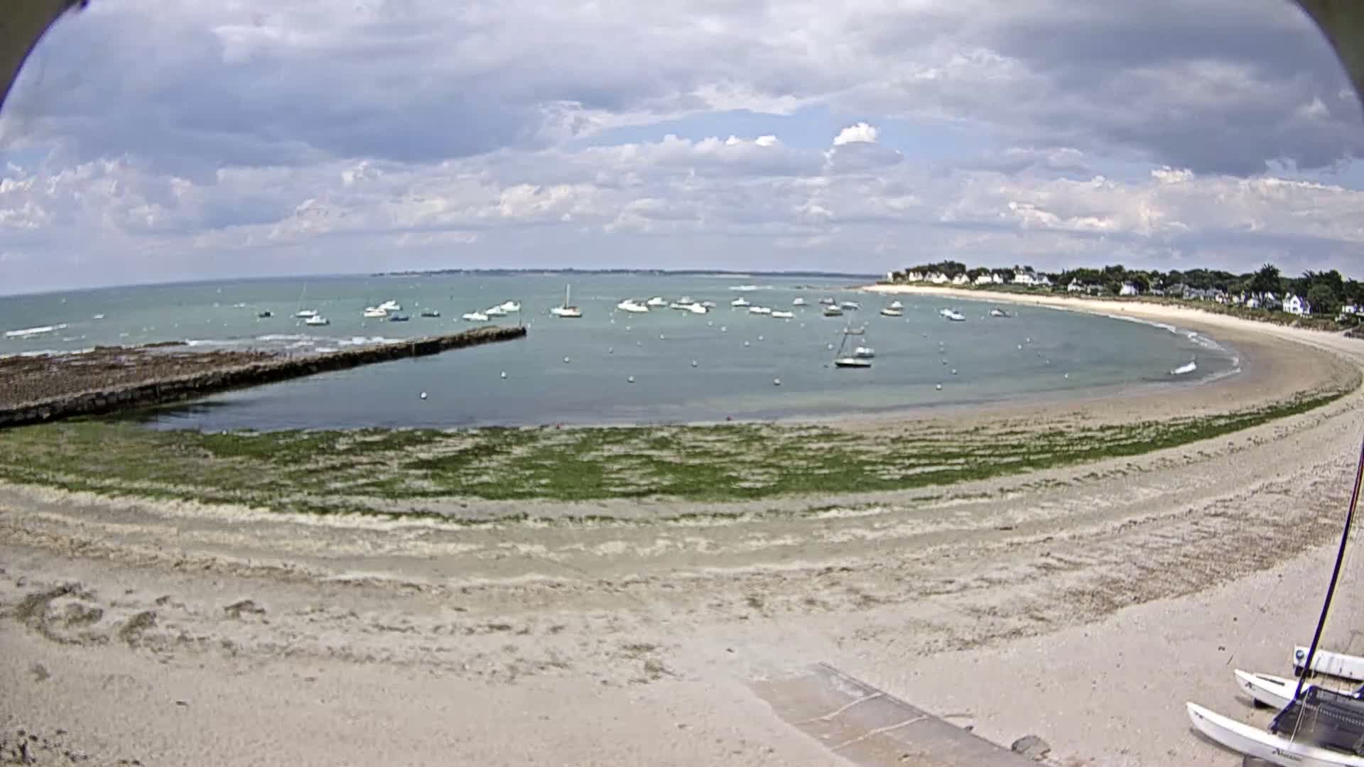A sandy beach curves around a calm bay filled with numerous small boats under a partly cloudy sky.