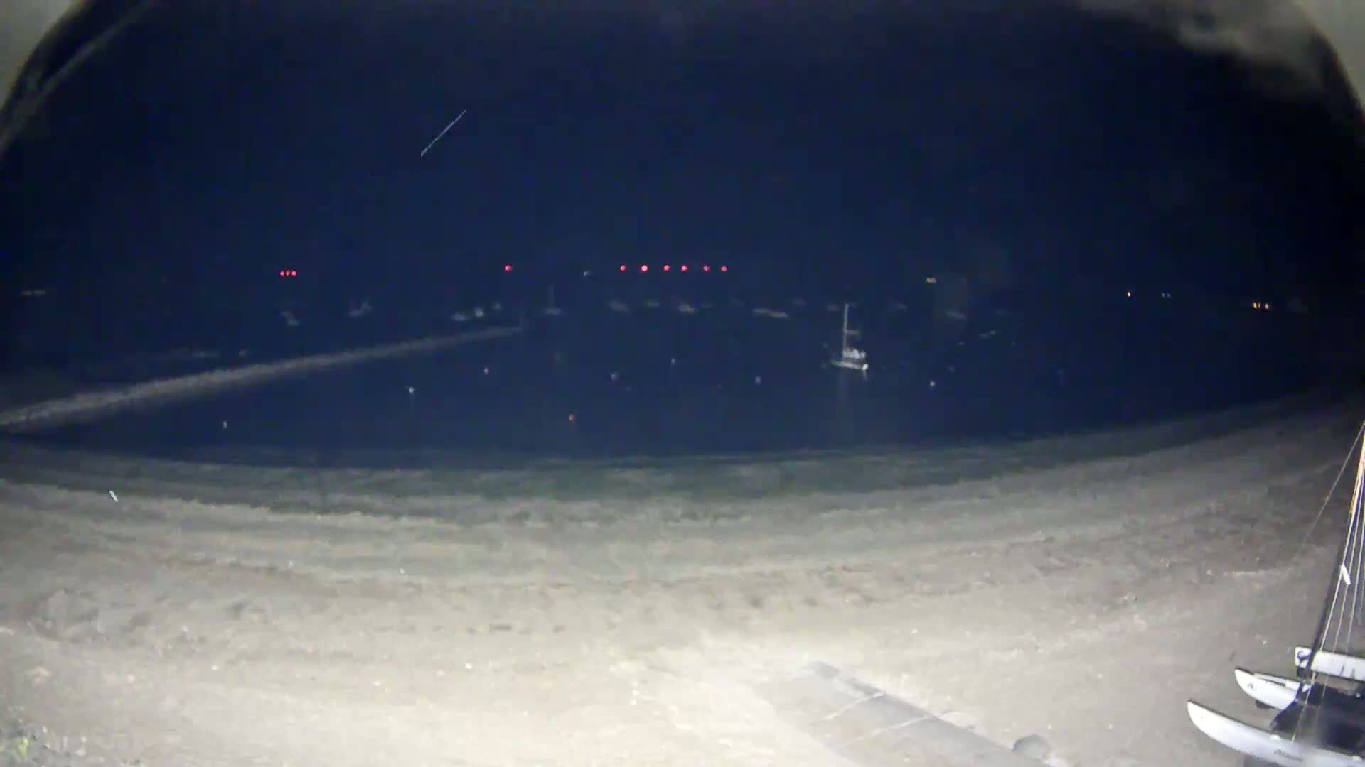 A nighttime view of a calm body of water with boats moored in a harbor, a sandy beach in the foreground, and two catamarans on the sand.
