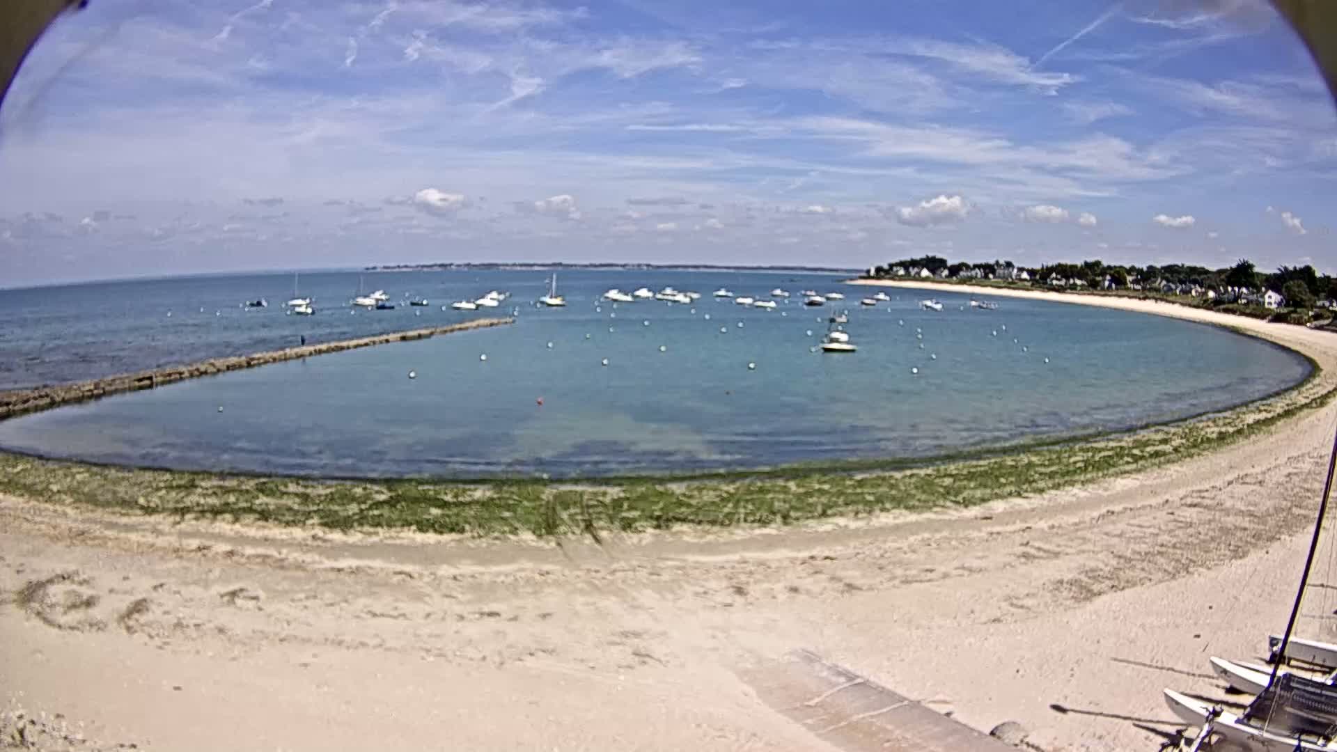 A sandy beach curves around a calm, clear bay dotted with numerous small boats under a mostly sunny sky.