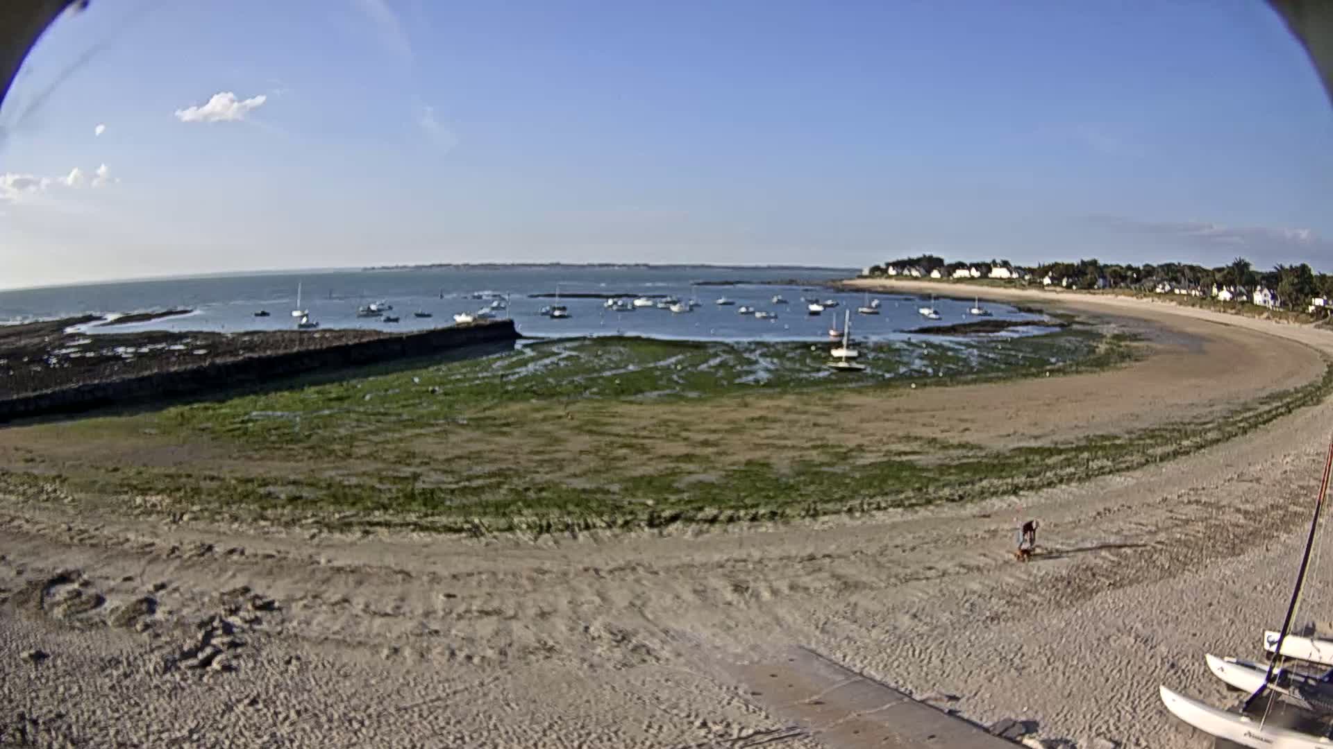 A sandy beach curves around a calm bay with numerous small boats at anchor under a mostly clear sky.