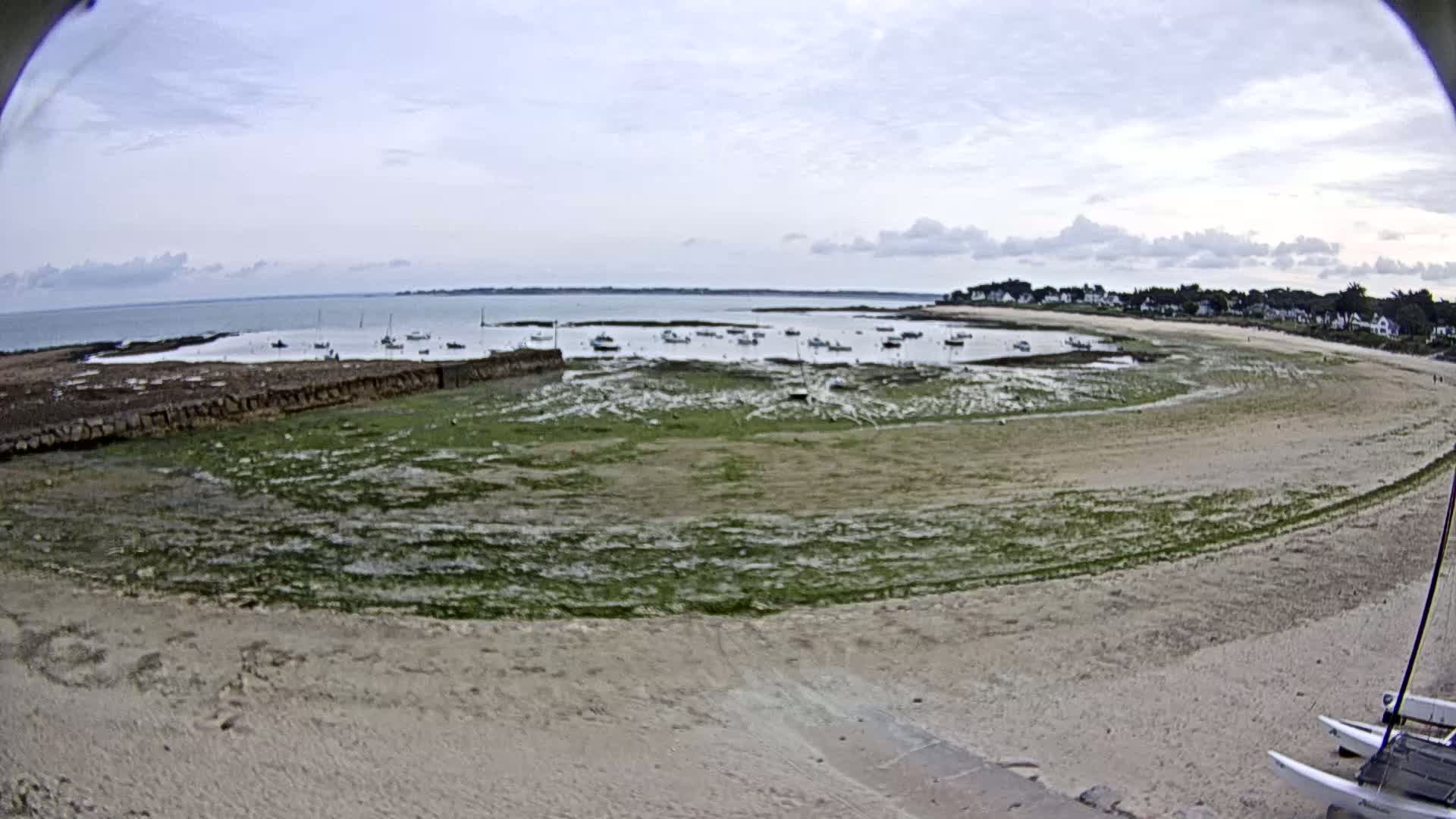 A sandy beach curves around a calm bay filled with small boats at low tide under a partly cloudy sky.
