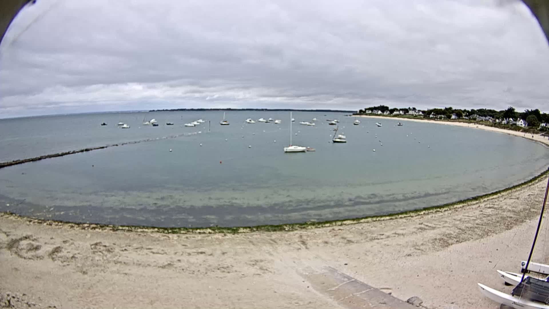 A sandy beach curves around a calm bay filled with numerous sailboats under a cloudy sky.