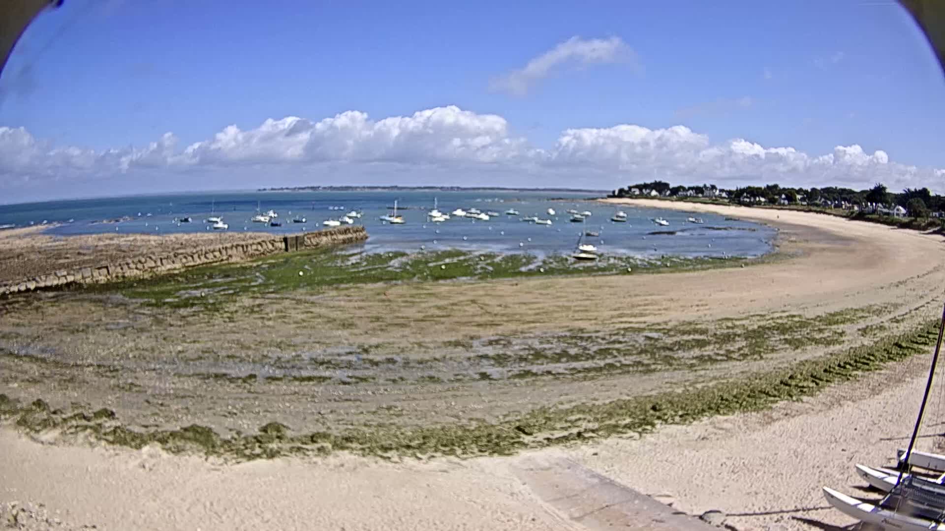 A sandy beach curves around a calm bay under a partly cloudy blue sky, with numerous small boats anchored in the water and several catamarans beached on the sand.