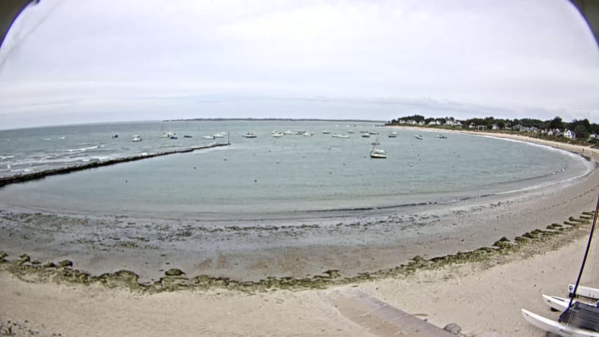 A calm, overcast day shows a sandy beach curving around a bay with numerous small boats moored in the water, a breakwater visible in the foreground.