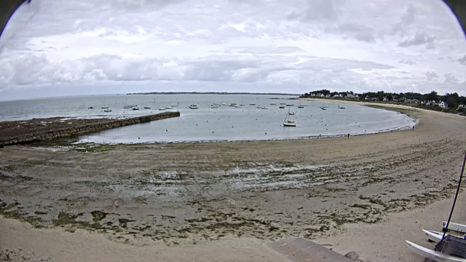 A sandy beach curves around a calm bay dotted with sailboats under a cloudy sky.