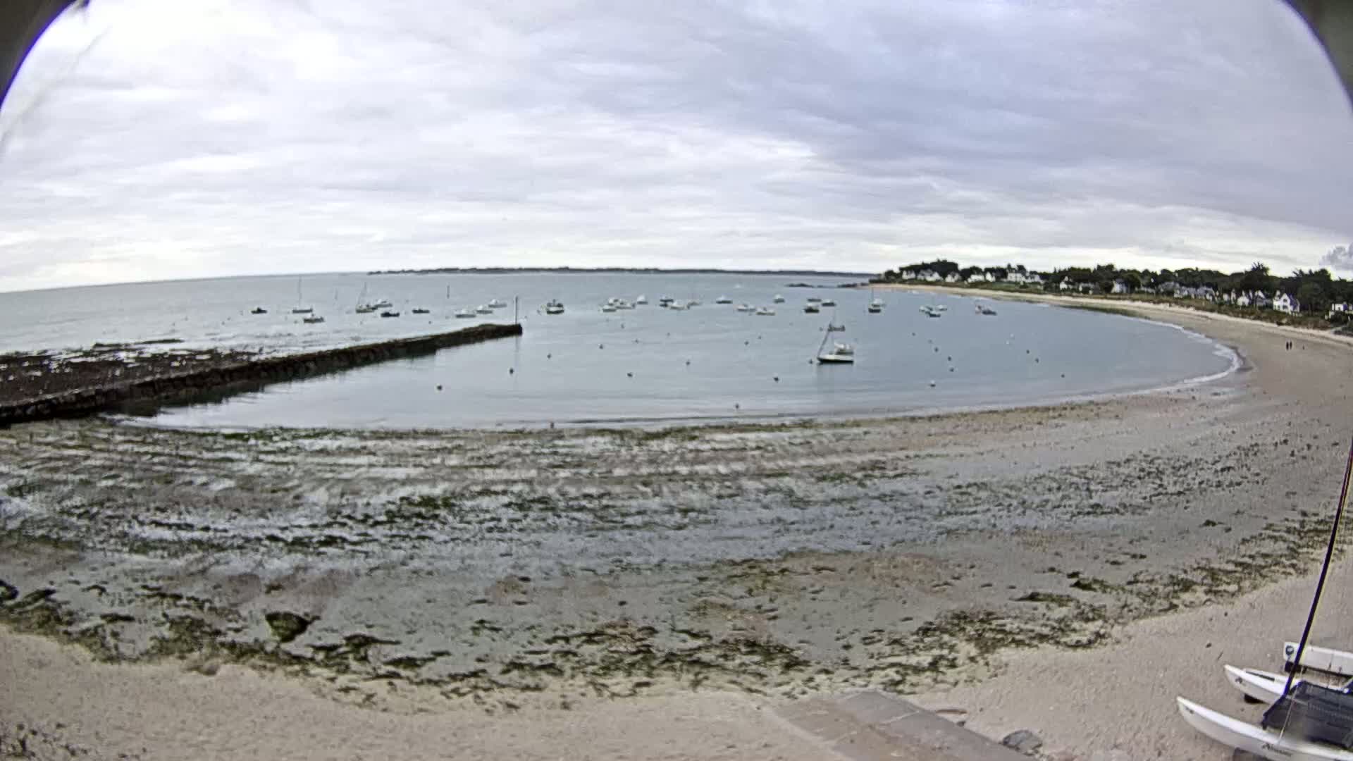 A calm, overcast day reveals a secluded bay with numerous small boats anchored in shallow, clear water, a sandy beach, and a low-lying stone wall extending into the sea.