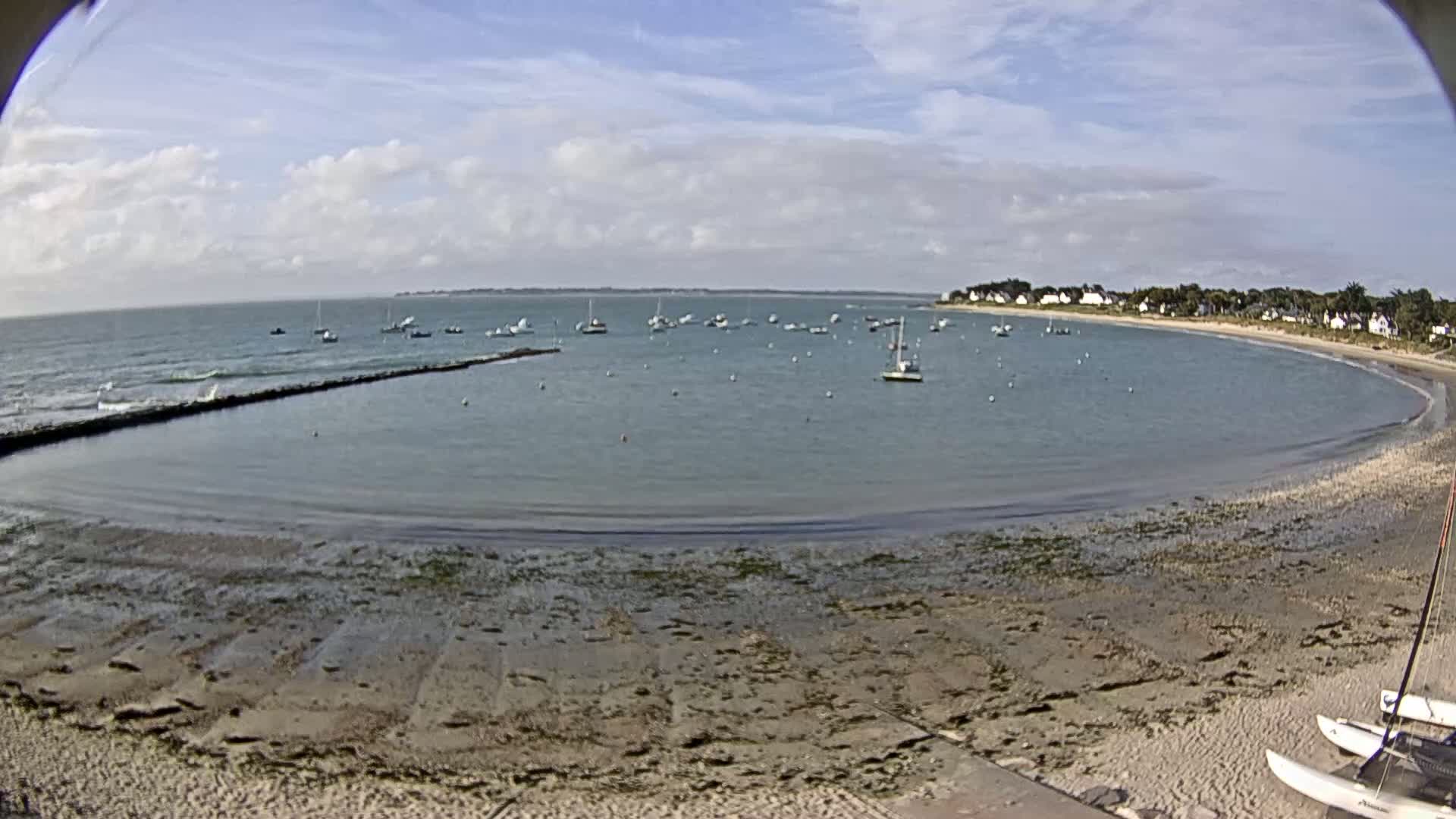 A calm, partly cloudy day reveals a secluded bay with numerous small sailboats anchored in shallow, clear water, a sandy beach, and a small residential area on the far shore.
