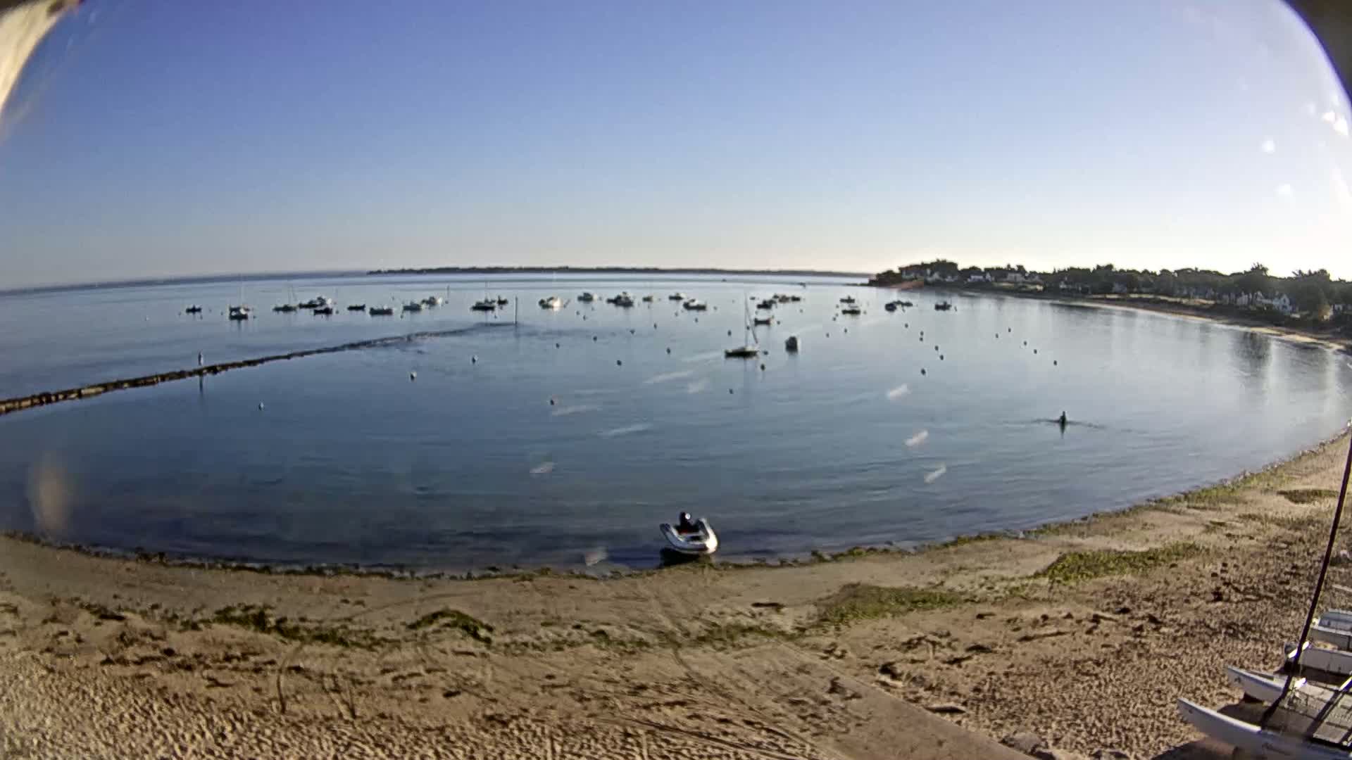 A sandy beach curves around a calm bay under a clear blue sky, with numerous small boats scattered across the water.