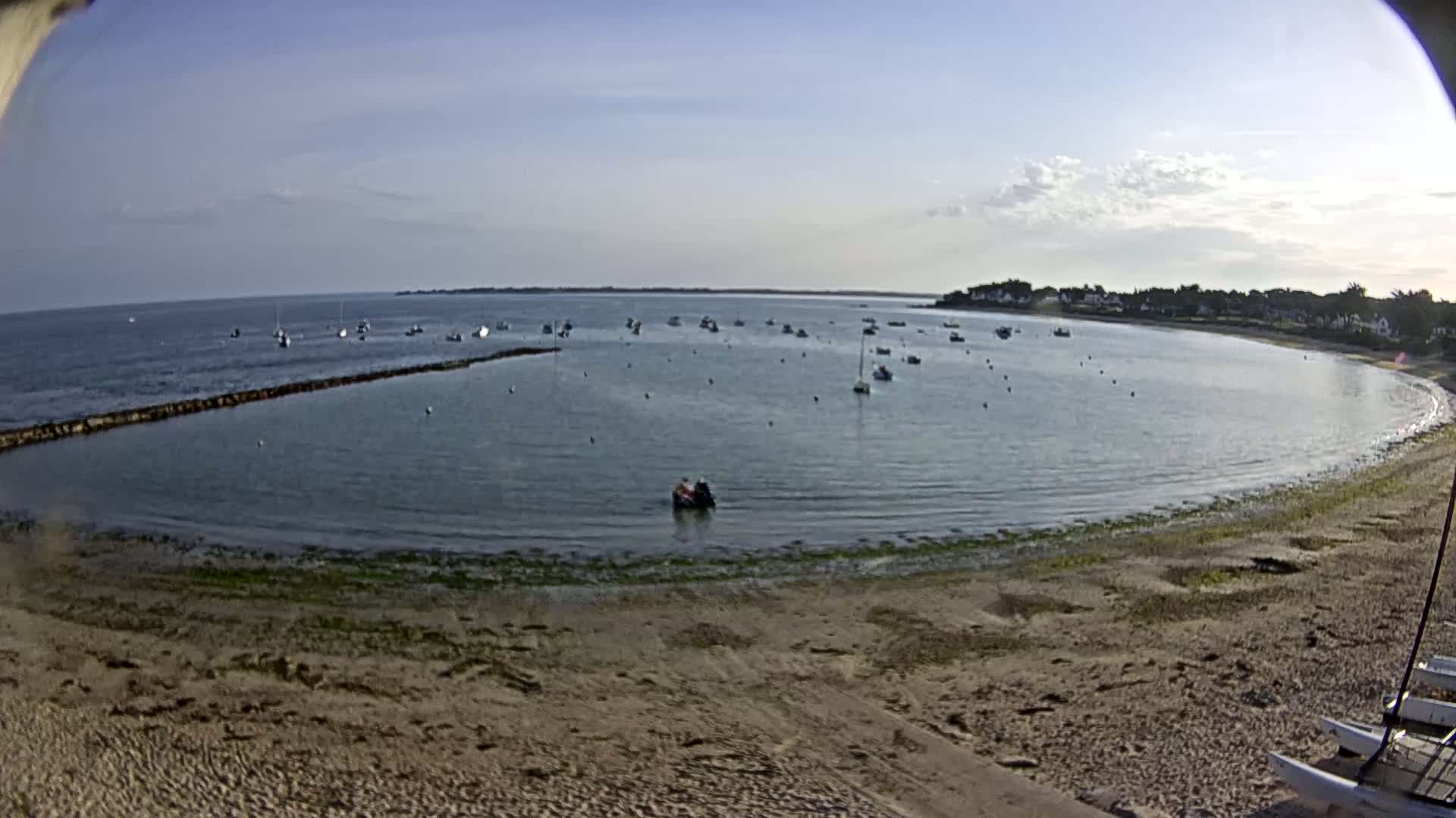 A sandy beach curves around a calm bay dotted with numerous sailboats under a mostly clear sky.