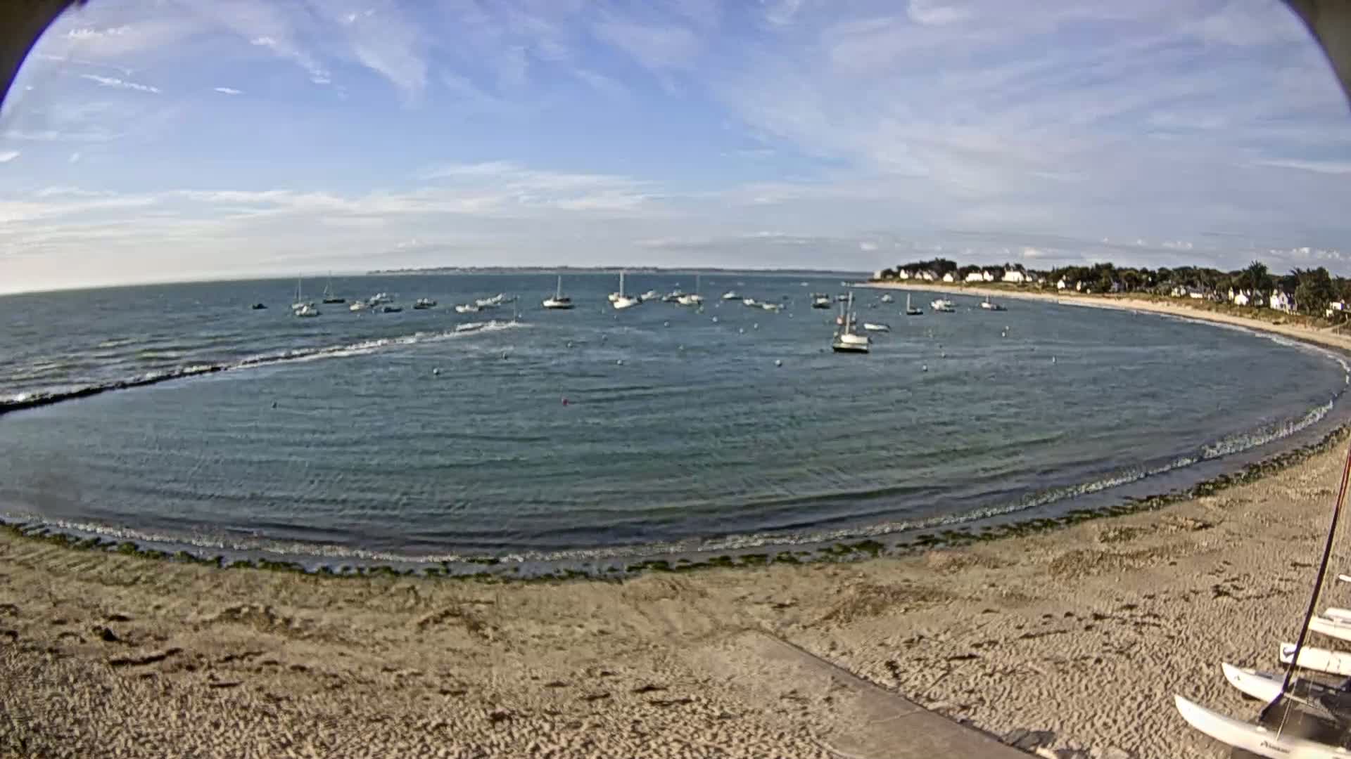 A sandy beach curves around a calm bay with numerous sailboats anchored in the water under a mostly sunny sky.