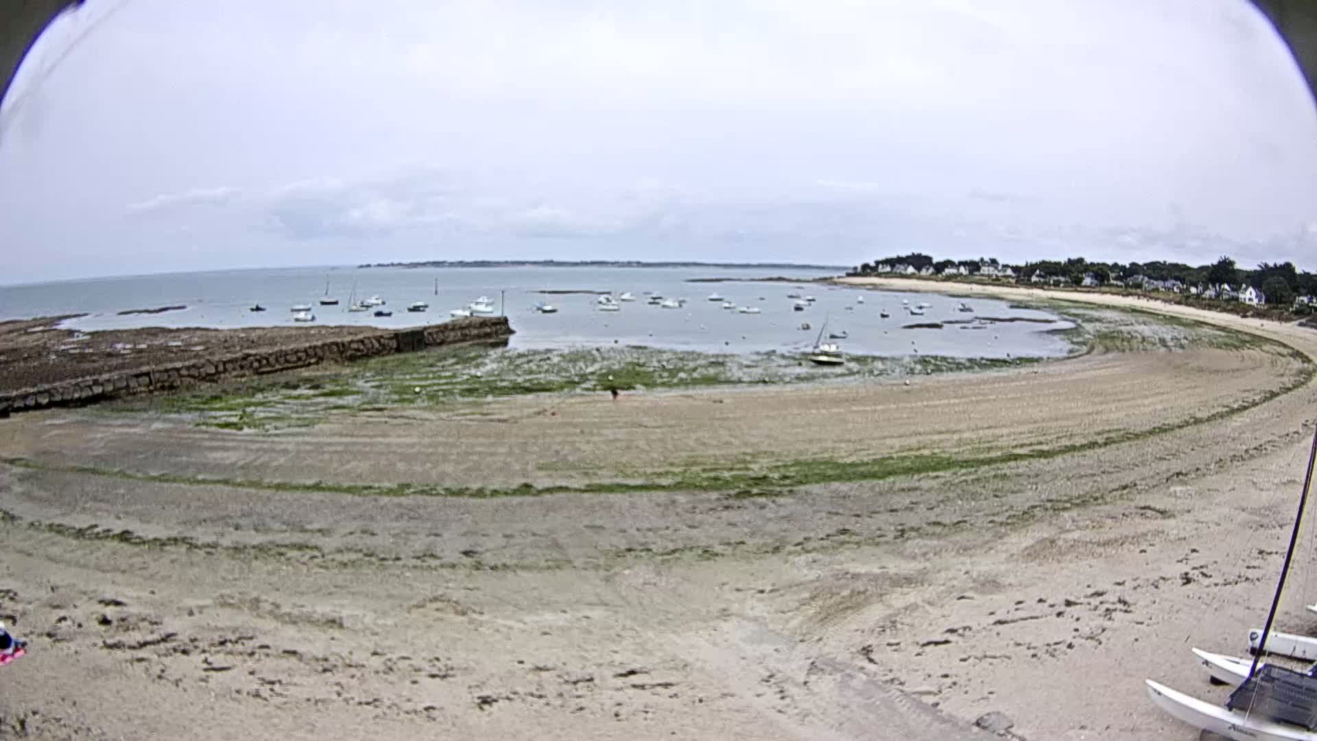 A sandy beach curves around a calm bay where several small boats are moored under an overcast sky.