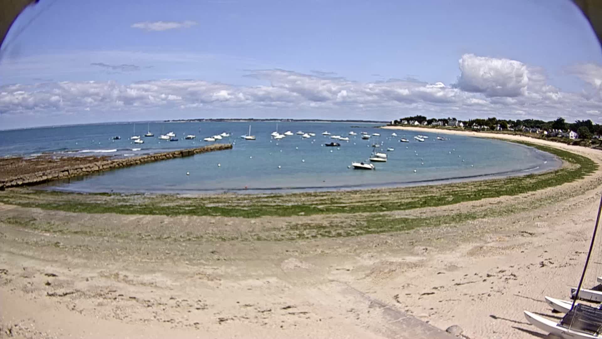 A sandy beach curves around a calm bay dotted with sailboats under a partly cloudy sky.