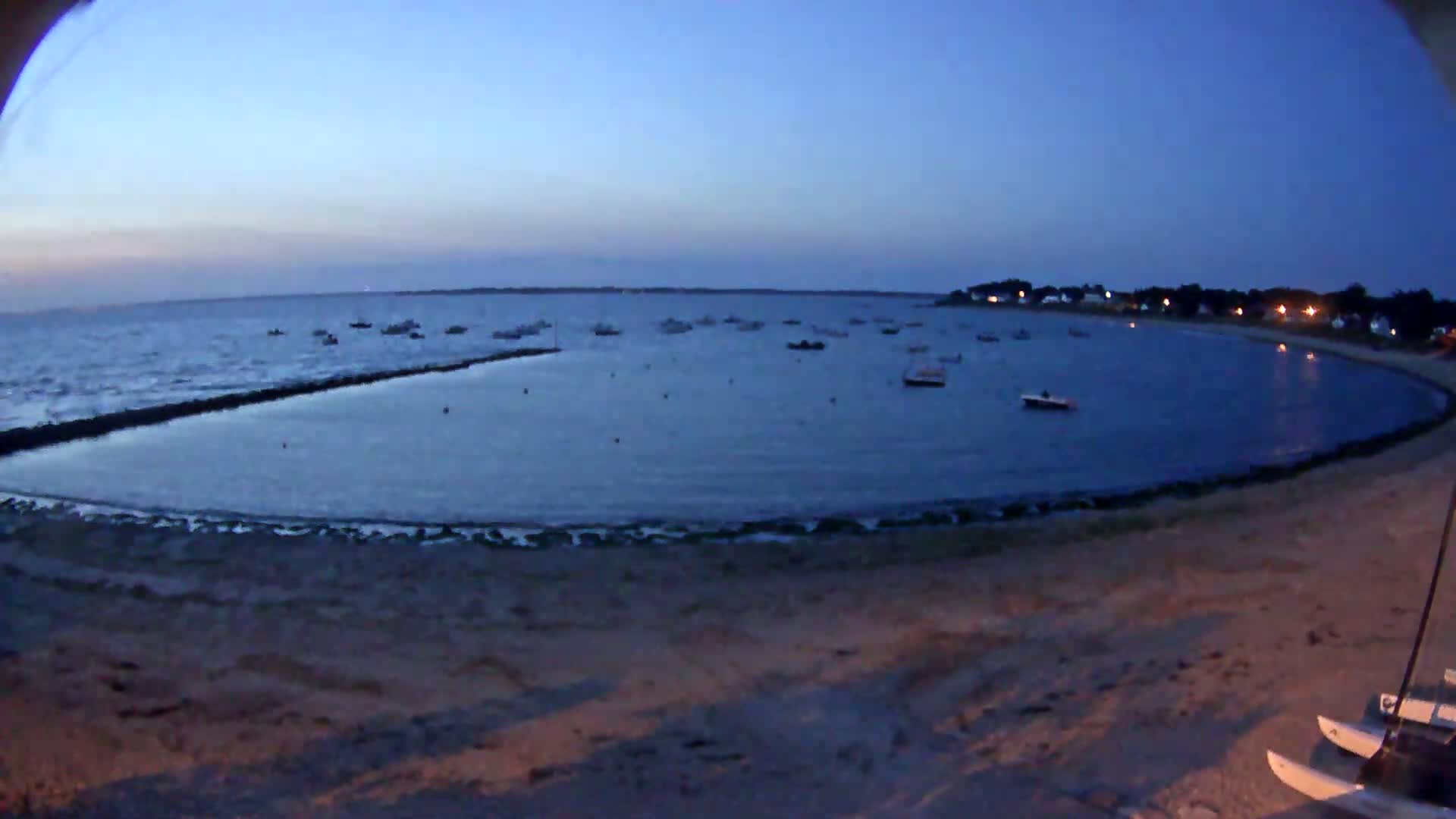A calm, dark-blue bay at twilight is dotted with numerous small boats near a sandy shore.